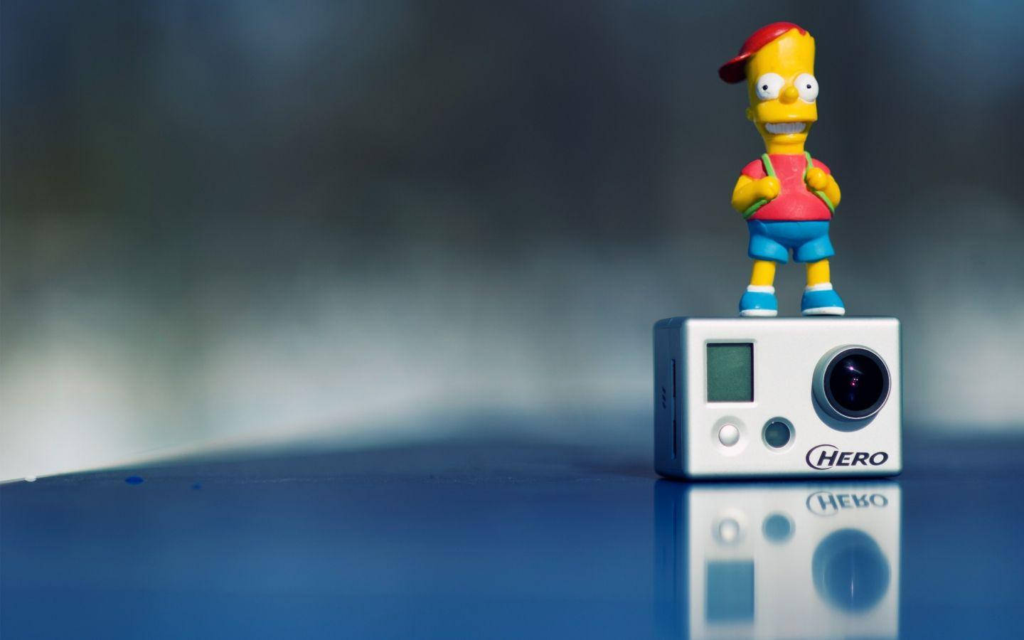 攝影 桌布 壁紙 — 一個巴特·辛普森公仔站在白色GoPro HERO相機上，位於反光藍色表面，背景模糊。