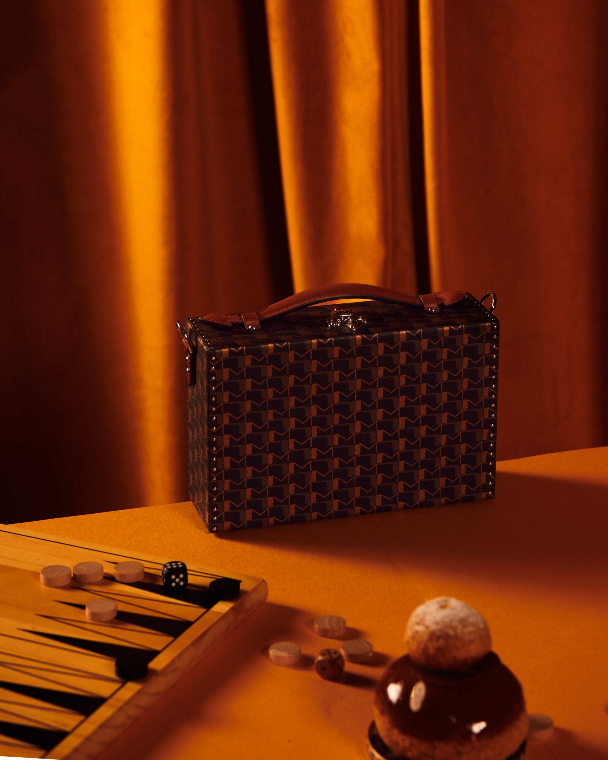 Luxury Aesthetic 桌布 壁紙 — 一個帶圖案的公文包放在桌子上，旁邊有一個雙陸棋套裝和一個巧克力甜點，在溫暖的金色燈光下。