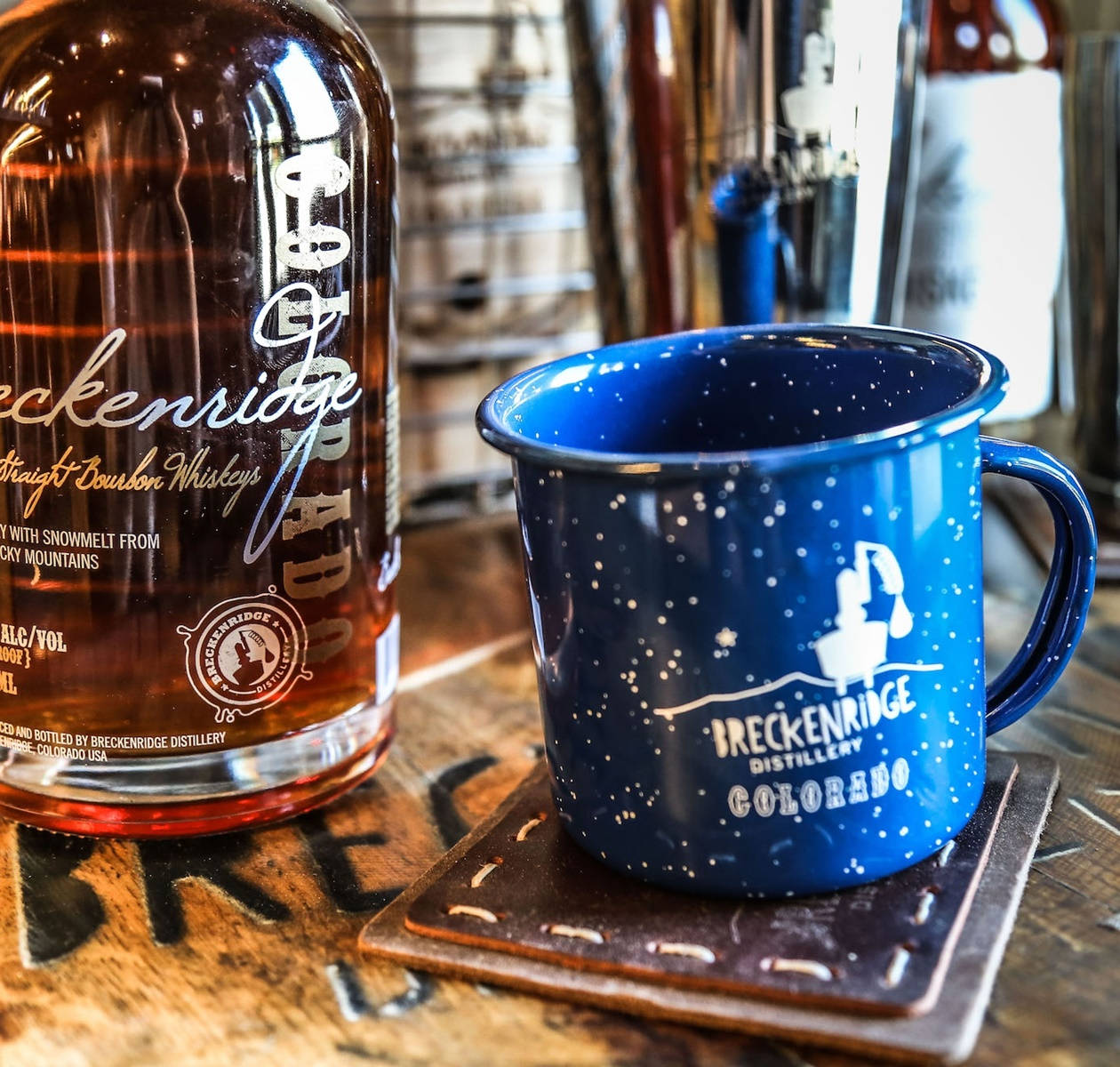 mug 桌布 壁紙 — 一個帶有 Breckenridge Distillery 標誌的藍色斑點馬克杯放在一瓶波本威士忌旁邊，置於鄉村風格的木製吧檯上。