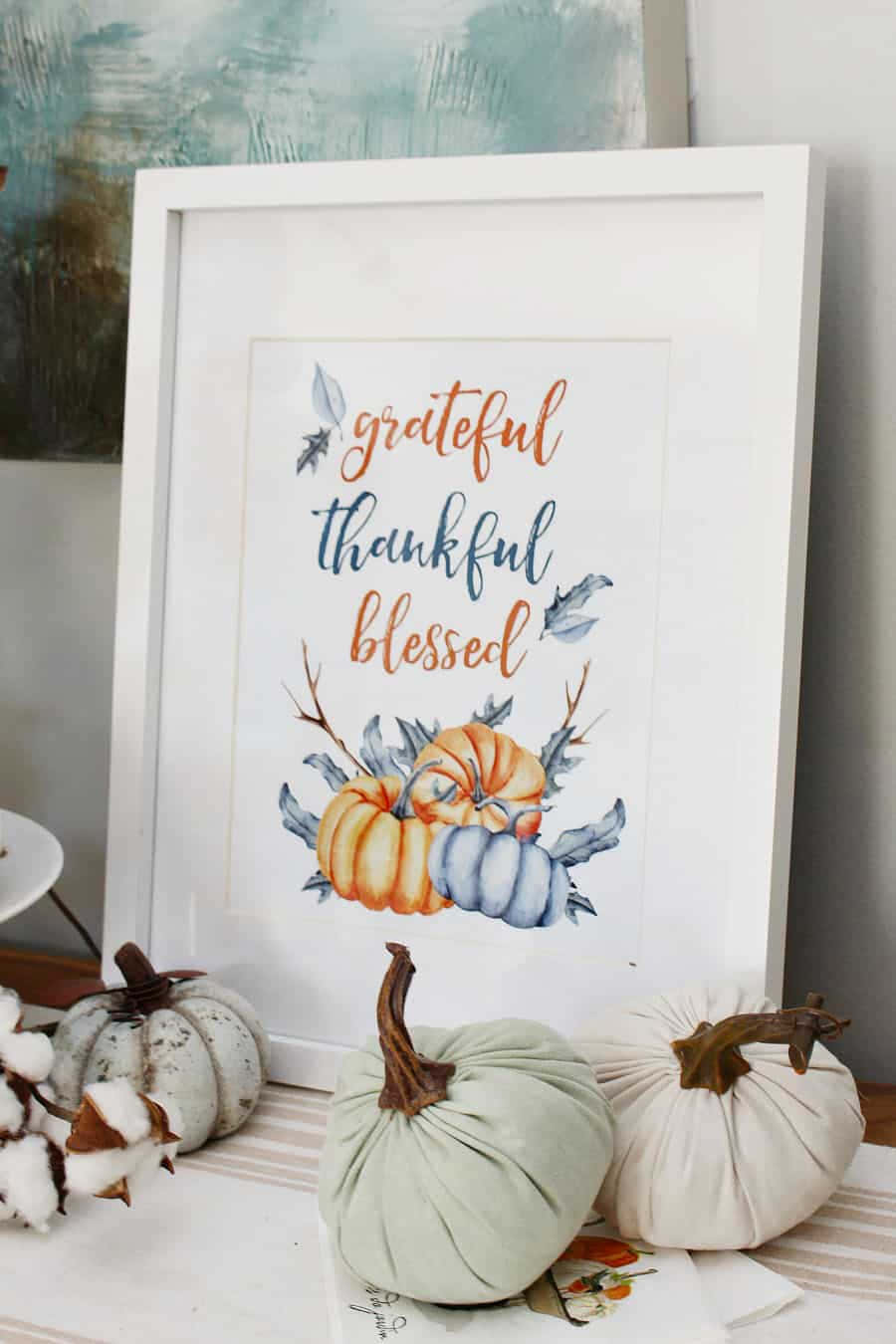 感恩節 桌布 壁紙 — 一個帶有 'grateful thankful blessed' 文字和南瓜的裱框水彩印刷品，旁邊有布料南瓜和棉花，放在條紋桌上。