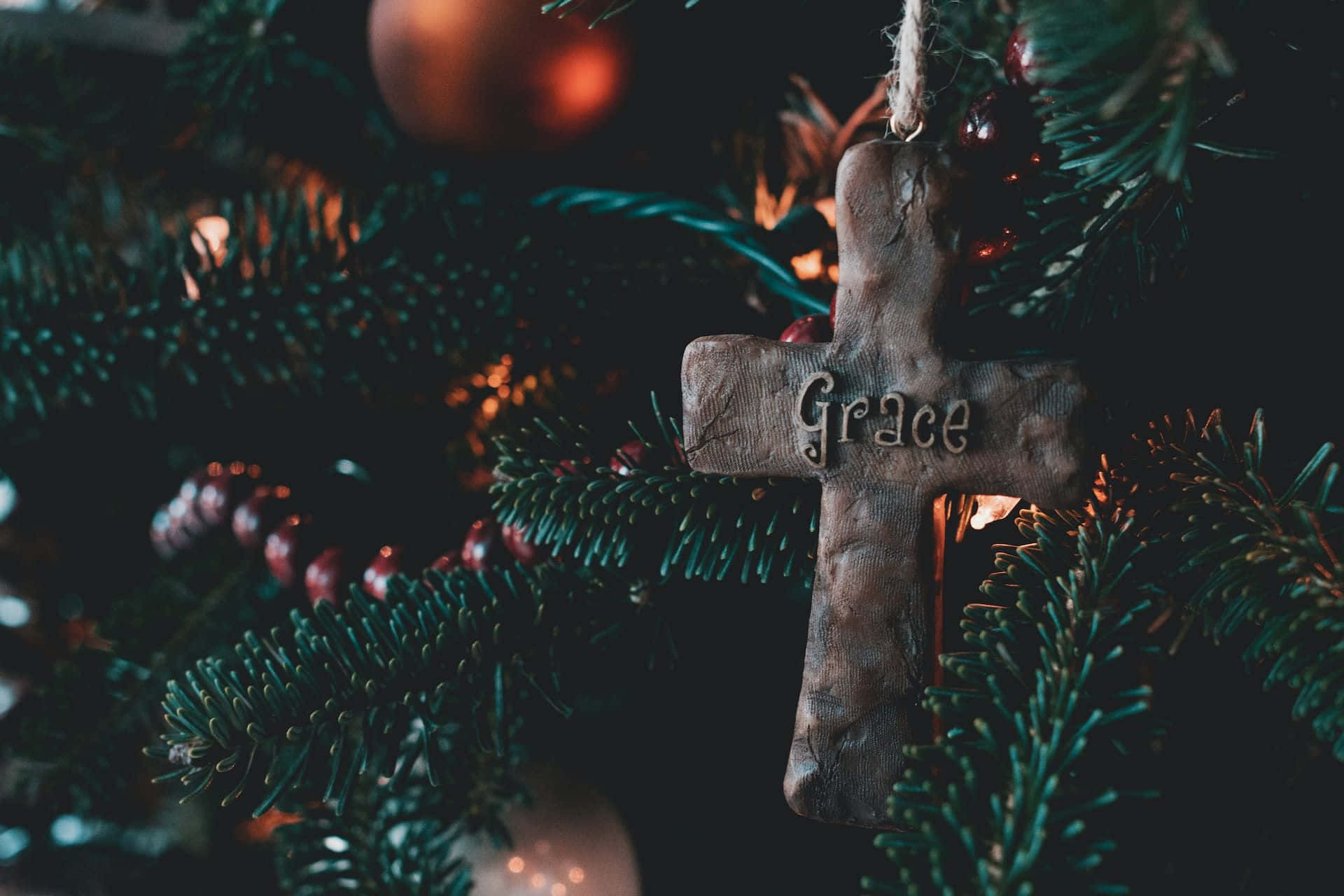 Religious Christmas 桌布 壁紙 — 一個帶有grace字樣的質樸木製十字架裝飾品，掛在聖誕樹上深綠色的松針和溫暖發光的燈光之間。
