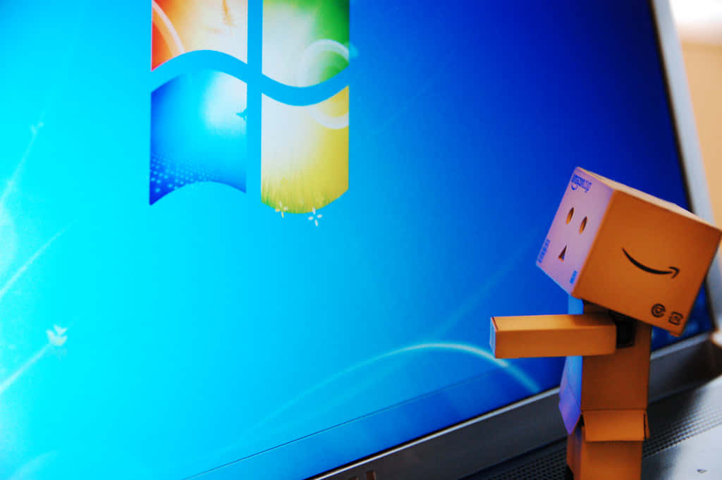 Cool Windows Desktop 桌布 壁紙 — 一個帶有亞馬遜標誌的紙板Danbo公仔在顯示Windows 7藍色桌面的筆記型電腦旁邊微笑。