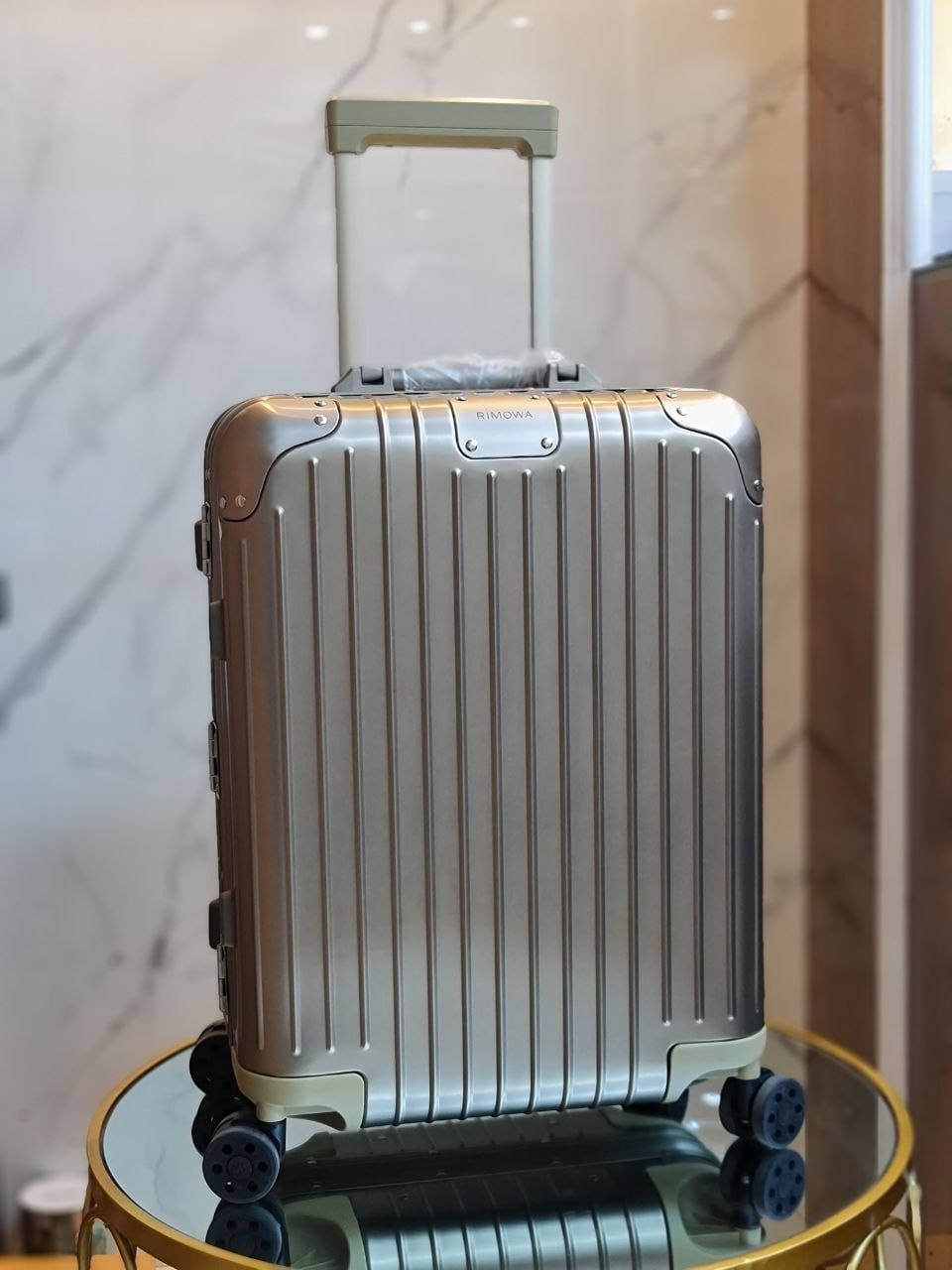 手機 桌布 壁紙 — 一個帶有垂直凹槽的時尚銀色Rimowa行李箱，直立放在大理石牆前的玻璃桌上。