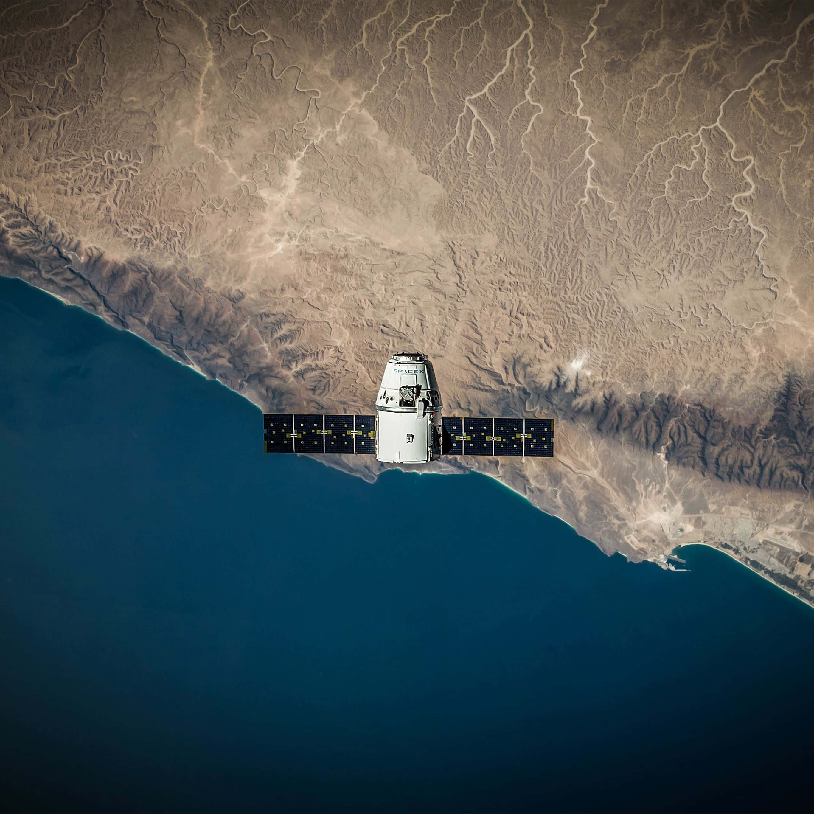 Satellite 桌布 壁紙 — 一個帶有太陽能板的白色衛星，在崎嶇的沙漠海岸與深藍色海洋交匯處上空運行。