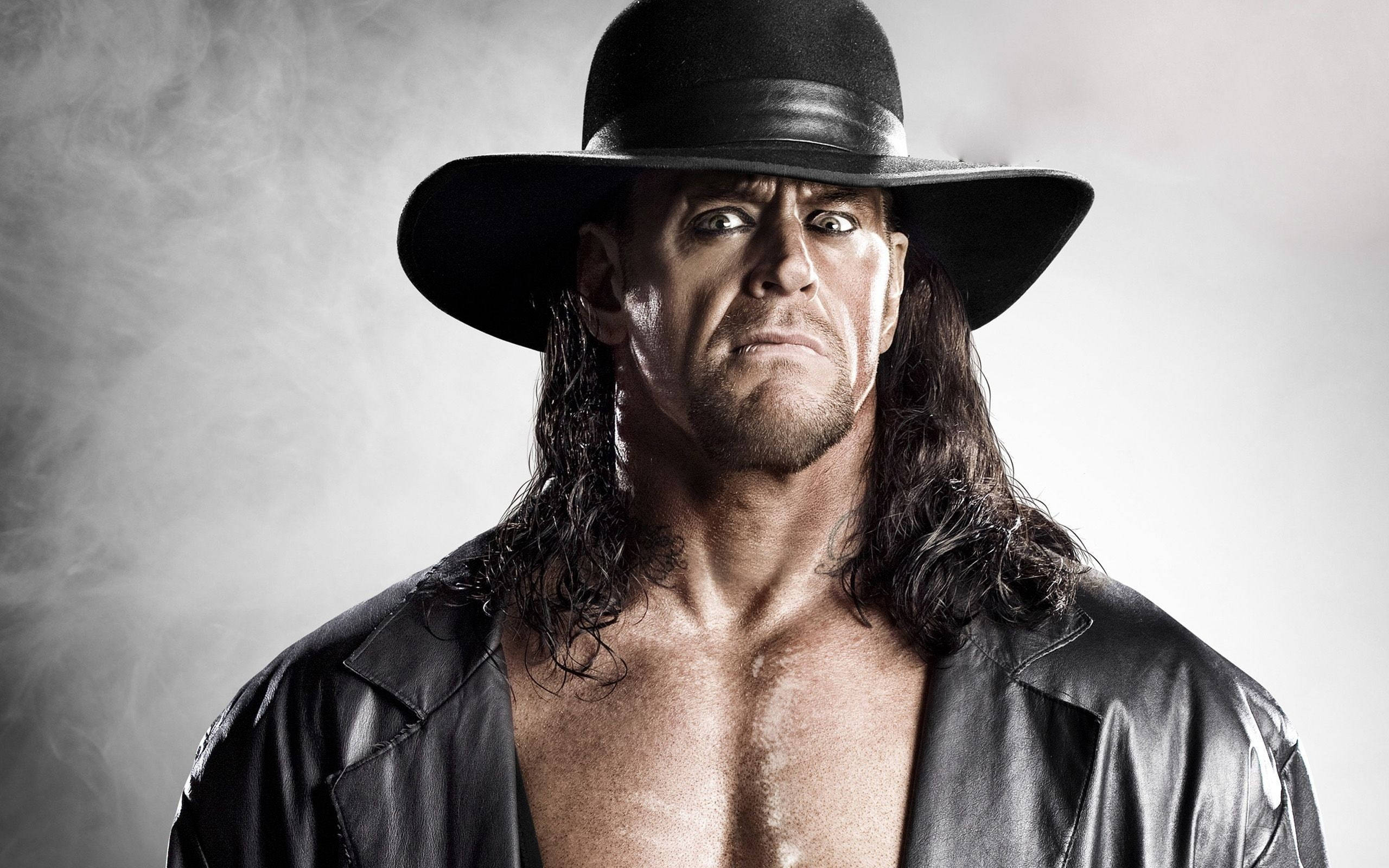 Undertaker 桌布 壁紙 — 一個帶有威脅性的摔角手，長長的黑髮和黑色帽子，在煙霧繚繞的灰色背景下凝視著。