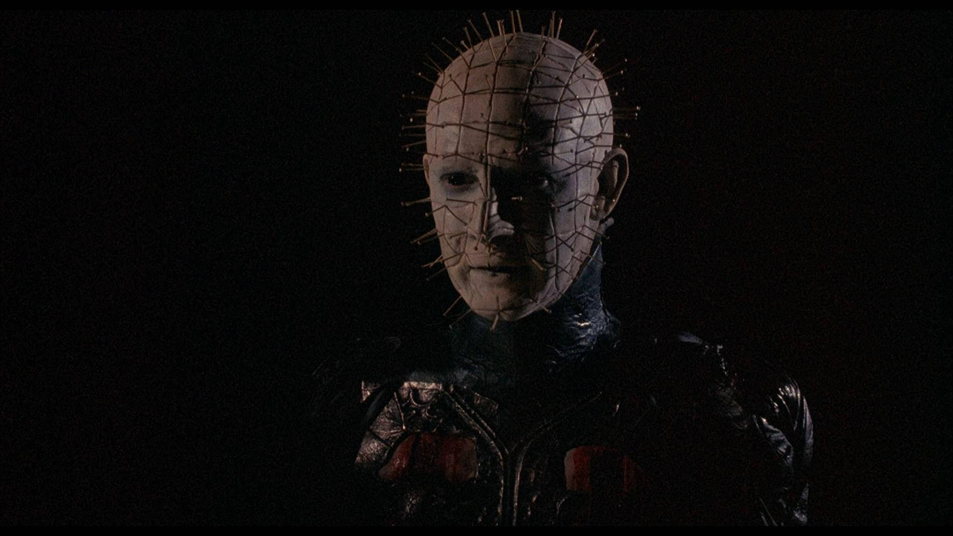 Pinhead 桌布 壁紙 — 一個帶有尖刺網格圖案頭部和深色皮革服裝的威脅性人物站在陰影黑暗中。