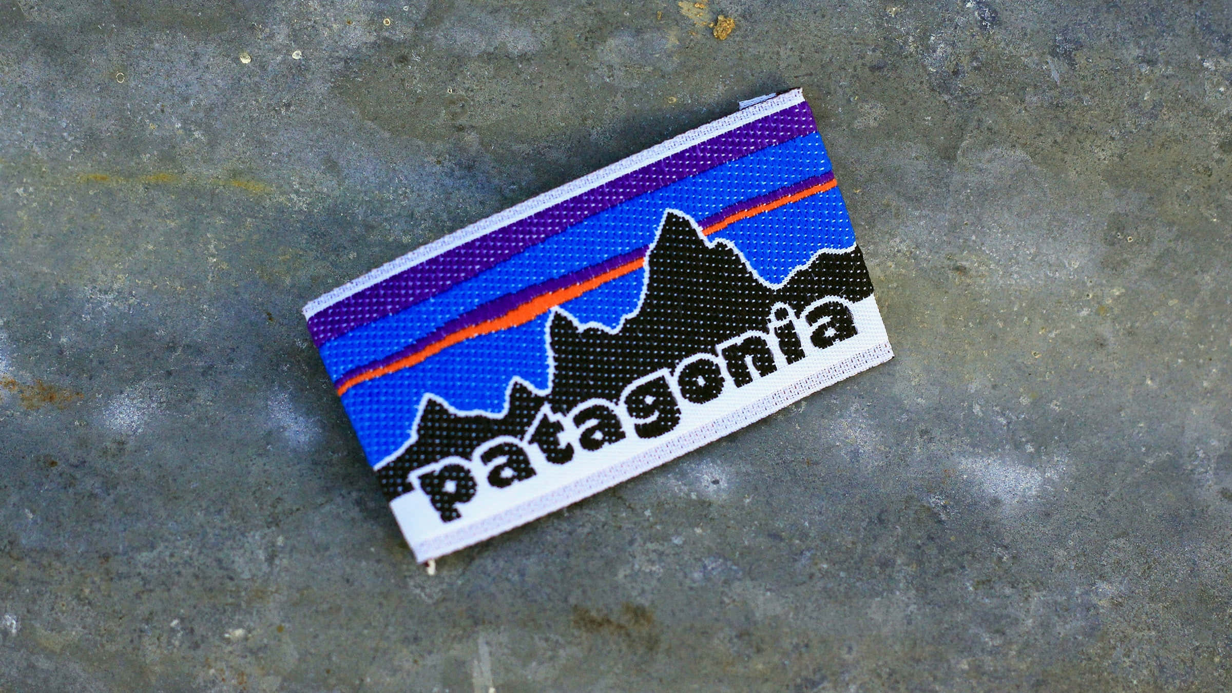 Patagonia 桌布 壁紙 — 一個帶有山脈標誌的Patagonia編織補丁放在有紋理的灰色混凝土表面上。