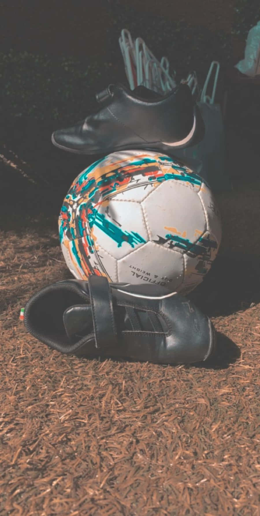 Soccer Ball 桌布 壁紙 — 一個帶有彩色油漆飛濺的足球放在黑色球靴之間，躺在昏暗燈光下的棕色覆蓋物上。