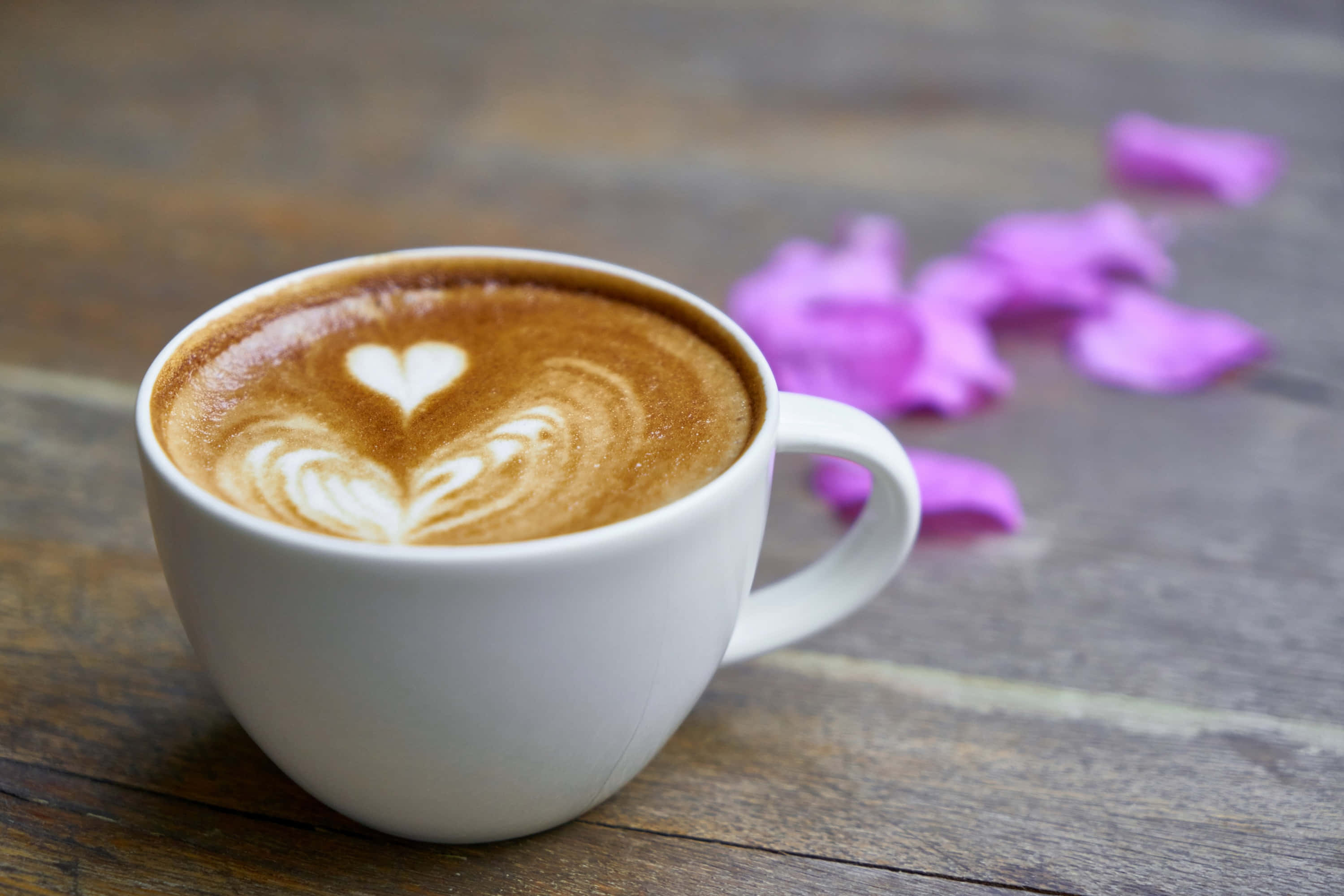Coffee Cup 桌布 壁紙 — 一個帶有心形拉花藝術的白色馬克杯，放在質樸的木桌上，旁邊散落著紫色花瓣。
