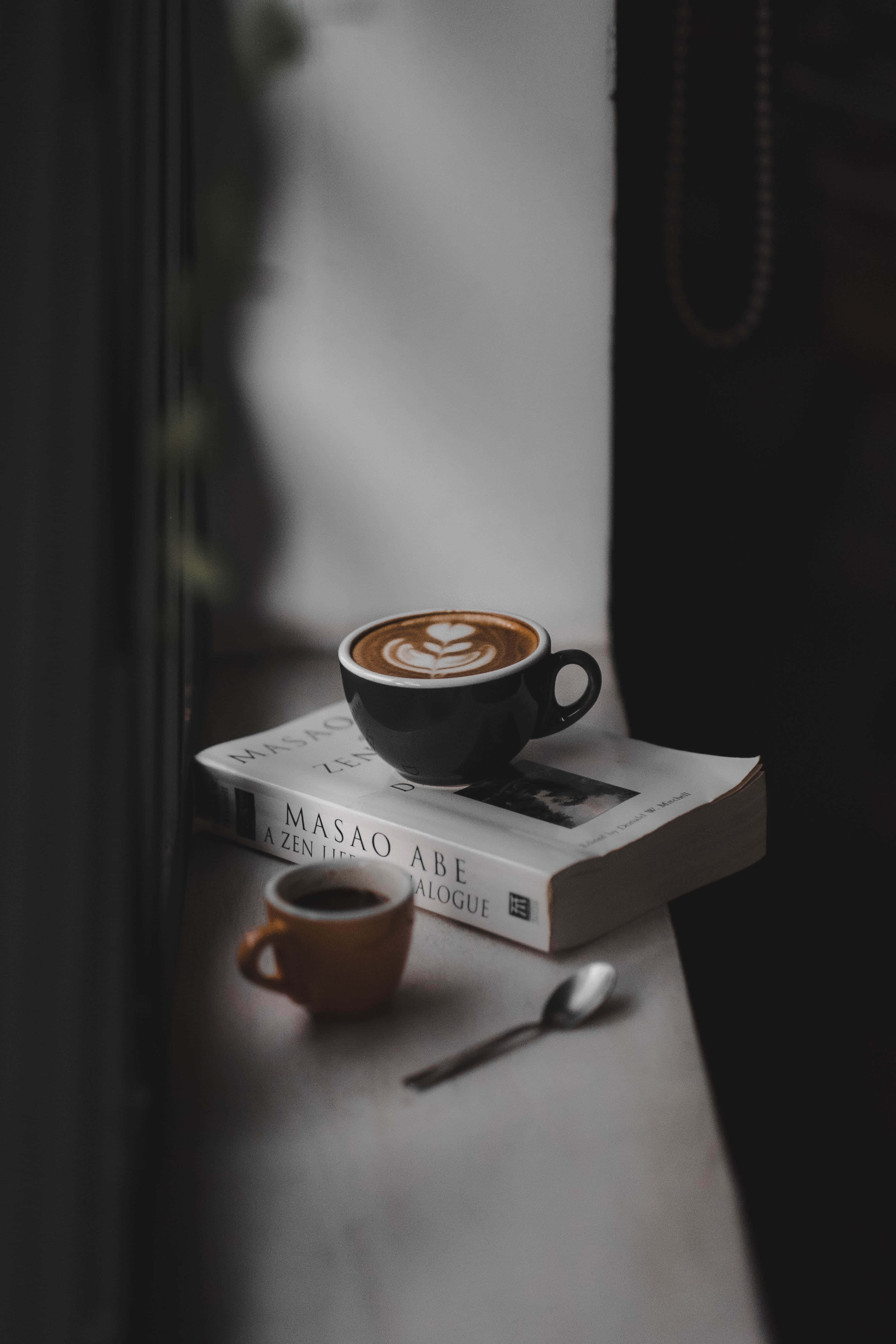 Coffee Aesthetic 桌布 壁紙 — 一個帶有心形拉花藝術的黑色咖啡杯放在大理石窗台上的一本禪書上，旁邊有一個棕色小杯和湯匙。