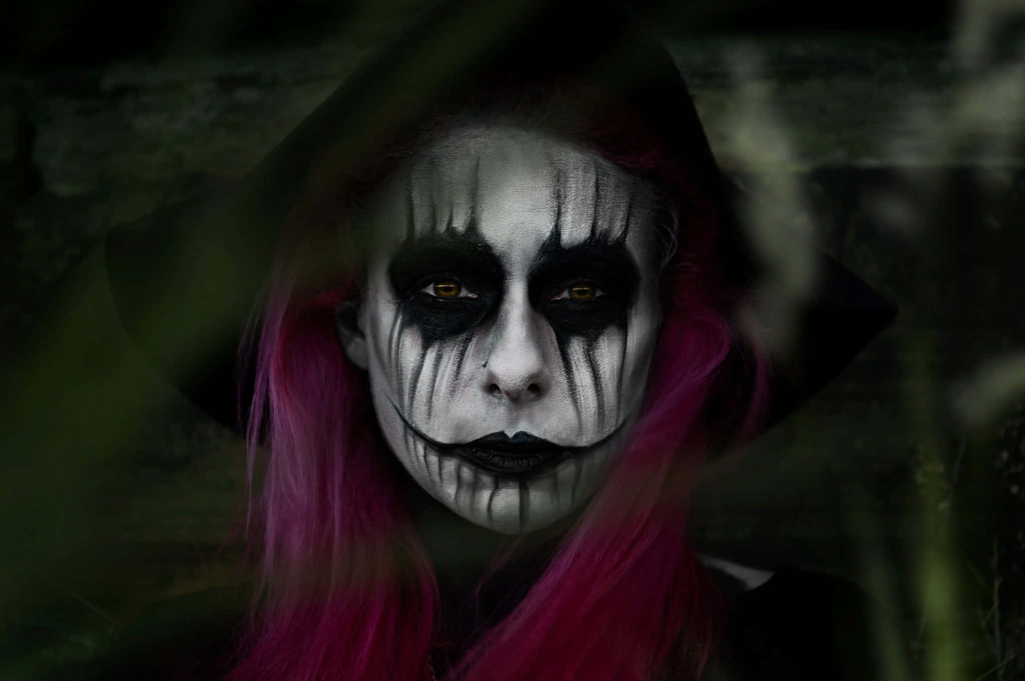 Halloween Makeup 桌布 壁紙 — 一個帶有戲劇性黑白臉部彩繪和鮮豔粉紅色頭髮的人，在黑暗、陰鬱的森林中透過樹葉窺視。