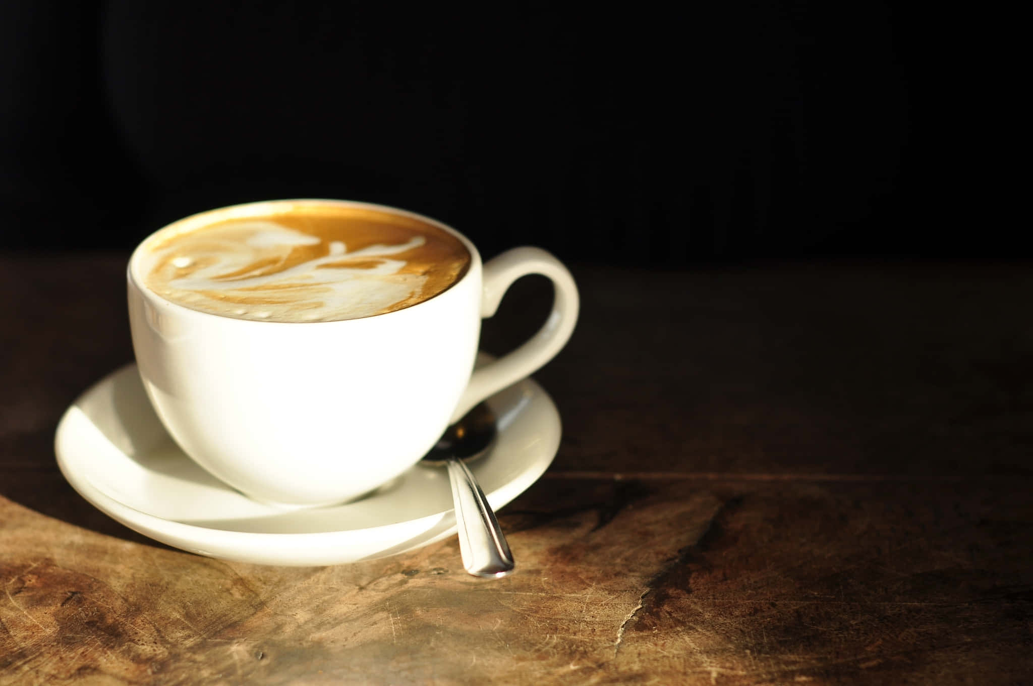 Coffee Aesthetic 桌布 壁紙 — 一個帶有拉花藝術的白色陶瓷咖啡杯放在碟子上，置於木桌上，沐浴在溫暖的陽光中，背景為深色。