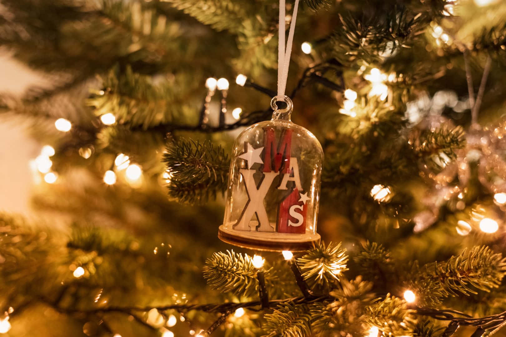 Teams Christmas 桌布 壁紙 — 一個帶有木製「MAS」字母的玻璃鈴鐺裝飾品掛在裝飾著溫暖閃爍仙女燈的松樹上。
