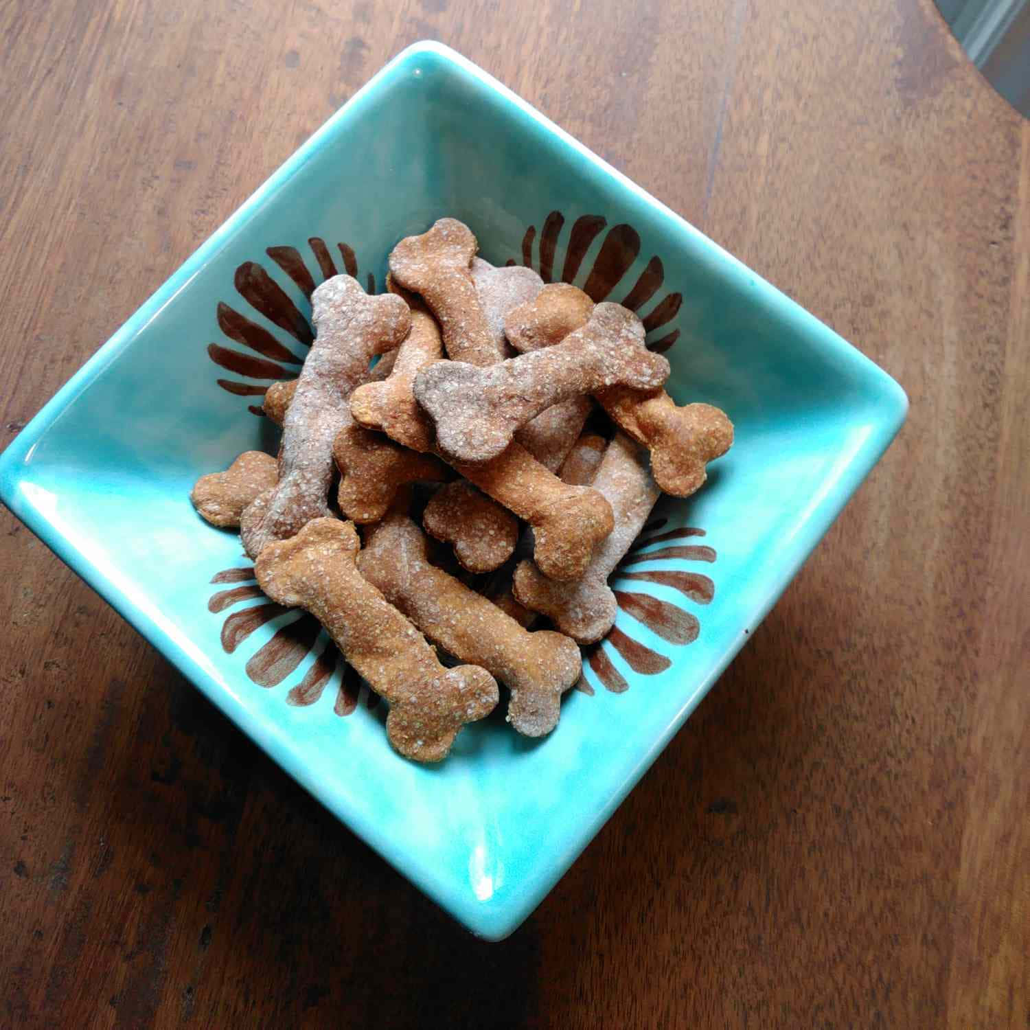 bones 桌布 壁紙 — 一個帶有棕色葉紋的綠松石方形碗，在木桌上盛放著骨形狗零食。