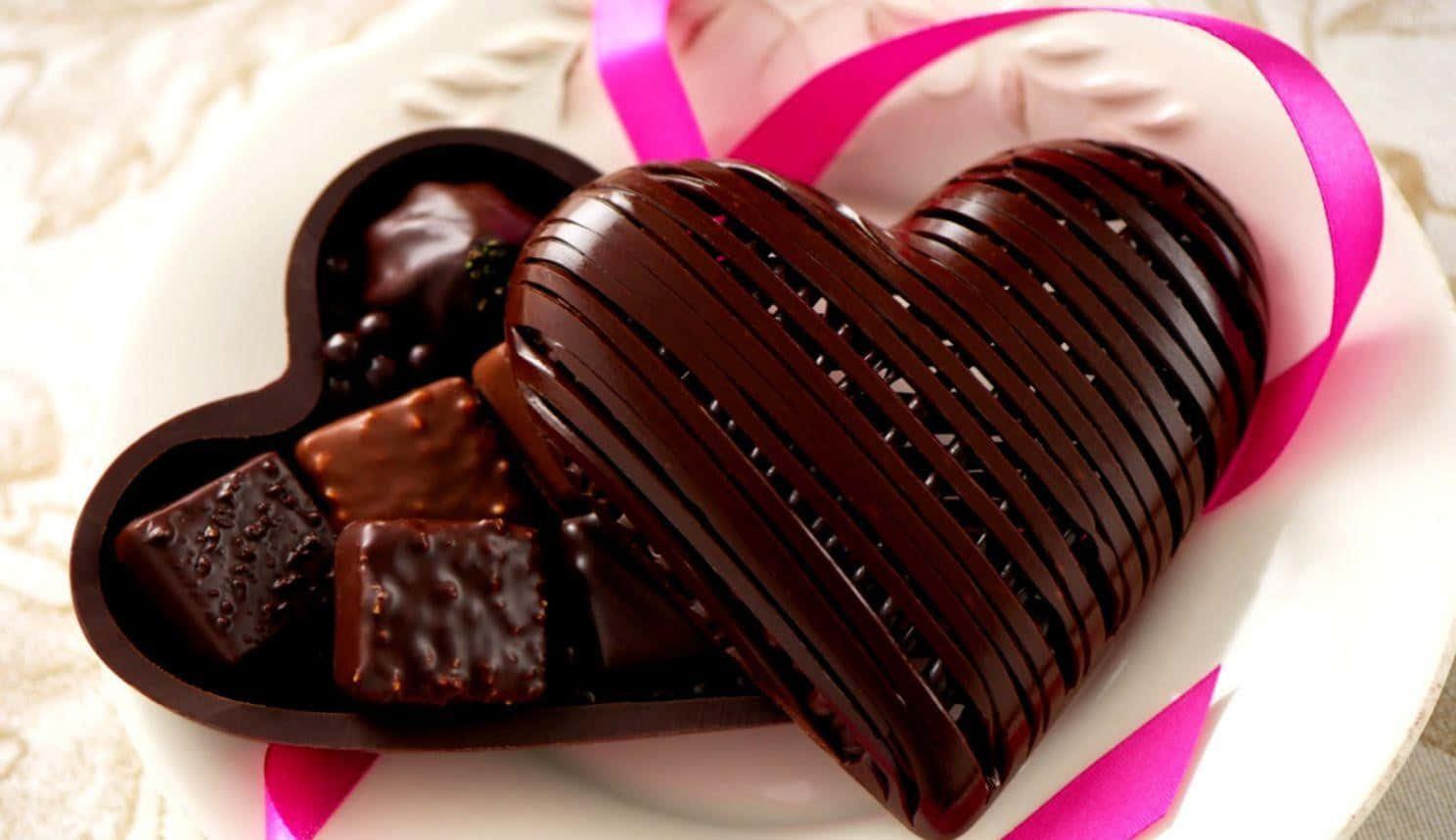 Chocolate 桌布 壁紙 — 一個帶有棱紋紋理的光澤心形巧克力放在白色盤子上，旁邊有各式巧克力和一條粉紅色絲帶。