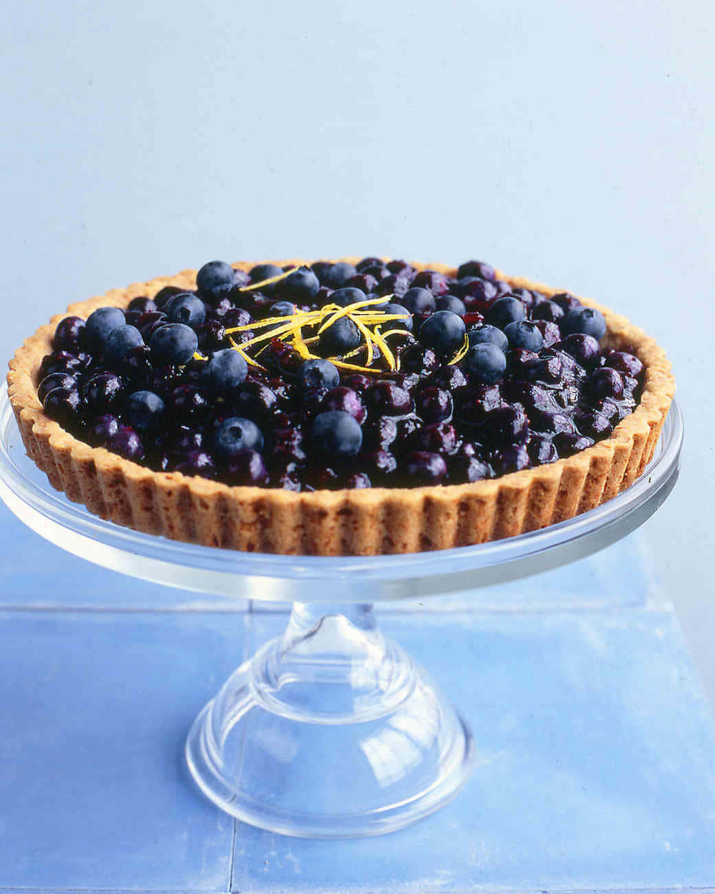 Blueberries Tart 桌布 壁紙 — 一個帶有檸檬皮的亮面藍莓塔放在玻璃底座上，背景是柔和的藍色。