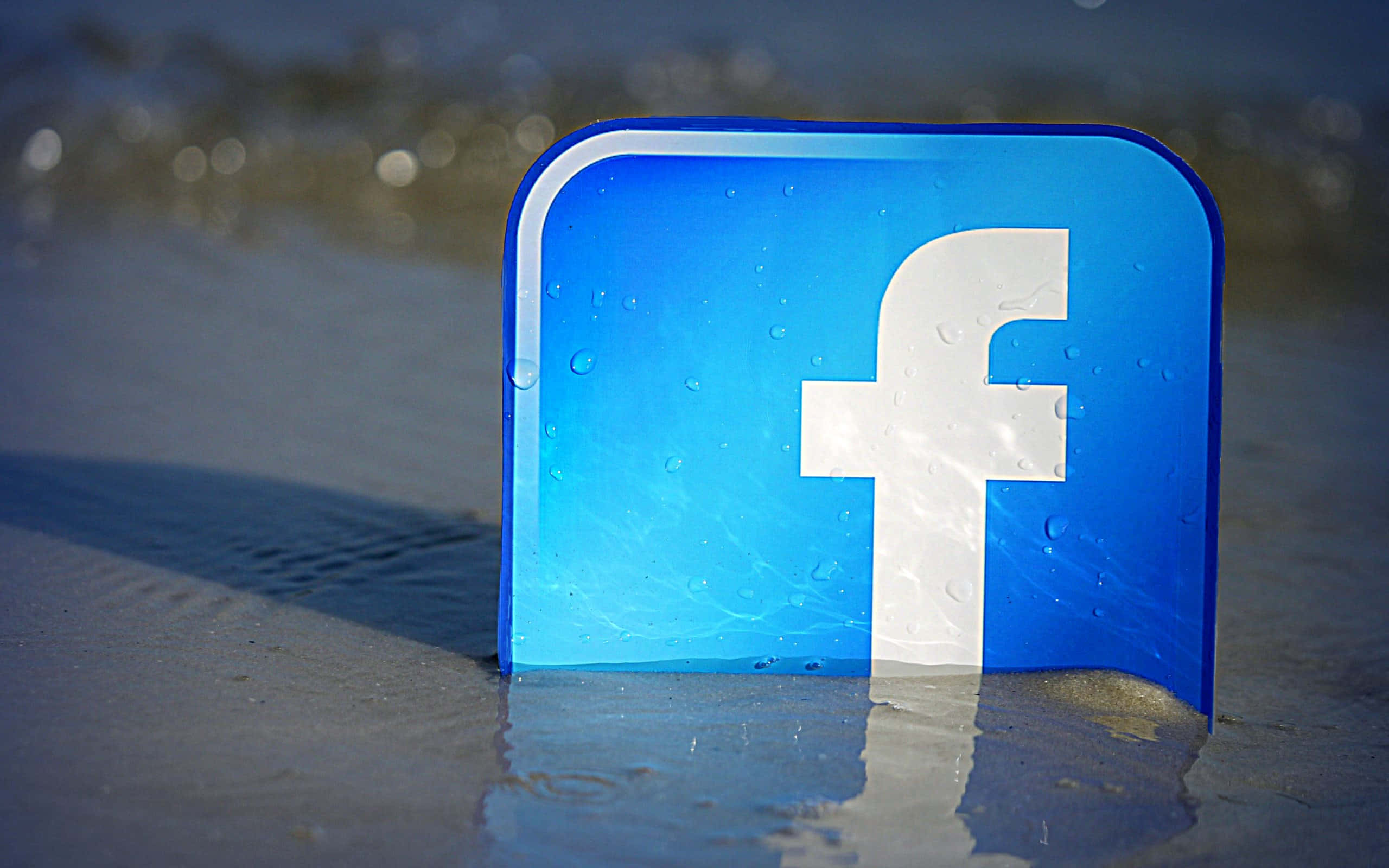 Facebook 桌布 壁紙 — 一個帶有水珠的藍色Facebook標誌牌矗立在沙灘邊緣的濕沙上，背景是模糊的海浪。