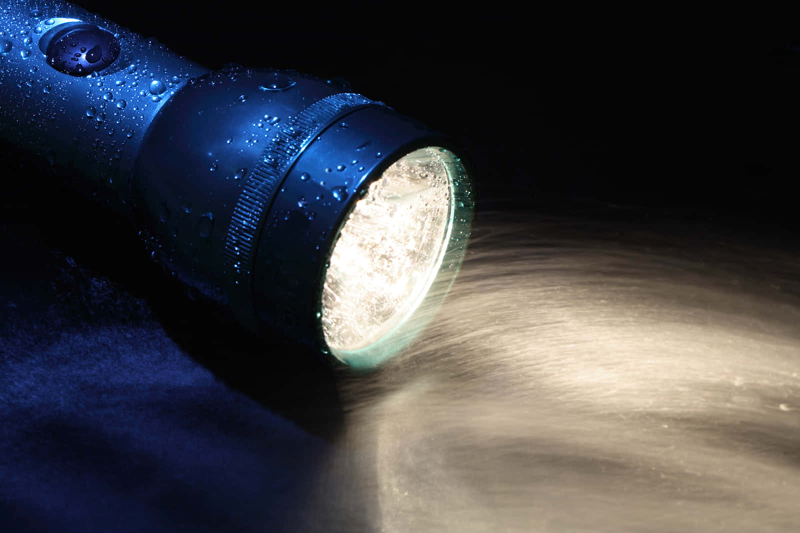 Flashlight 桌布 壁紙 — 一個帶有水珠的藍色手電筒在黑暗潮濕的表面上射出明亮的光芒，呈現戲劇性的特寫。