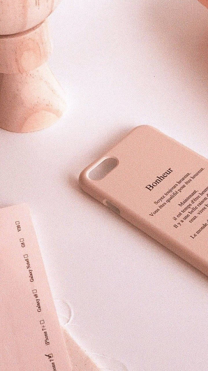Peach Aesthetic 桌布 壁紙 — 一個帶有法文文字的柔軟粉紅色手機殼放在一個極簡白色表面上的木燭台旁邊。