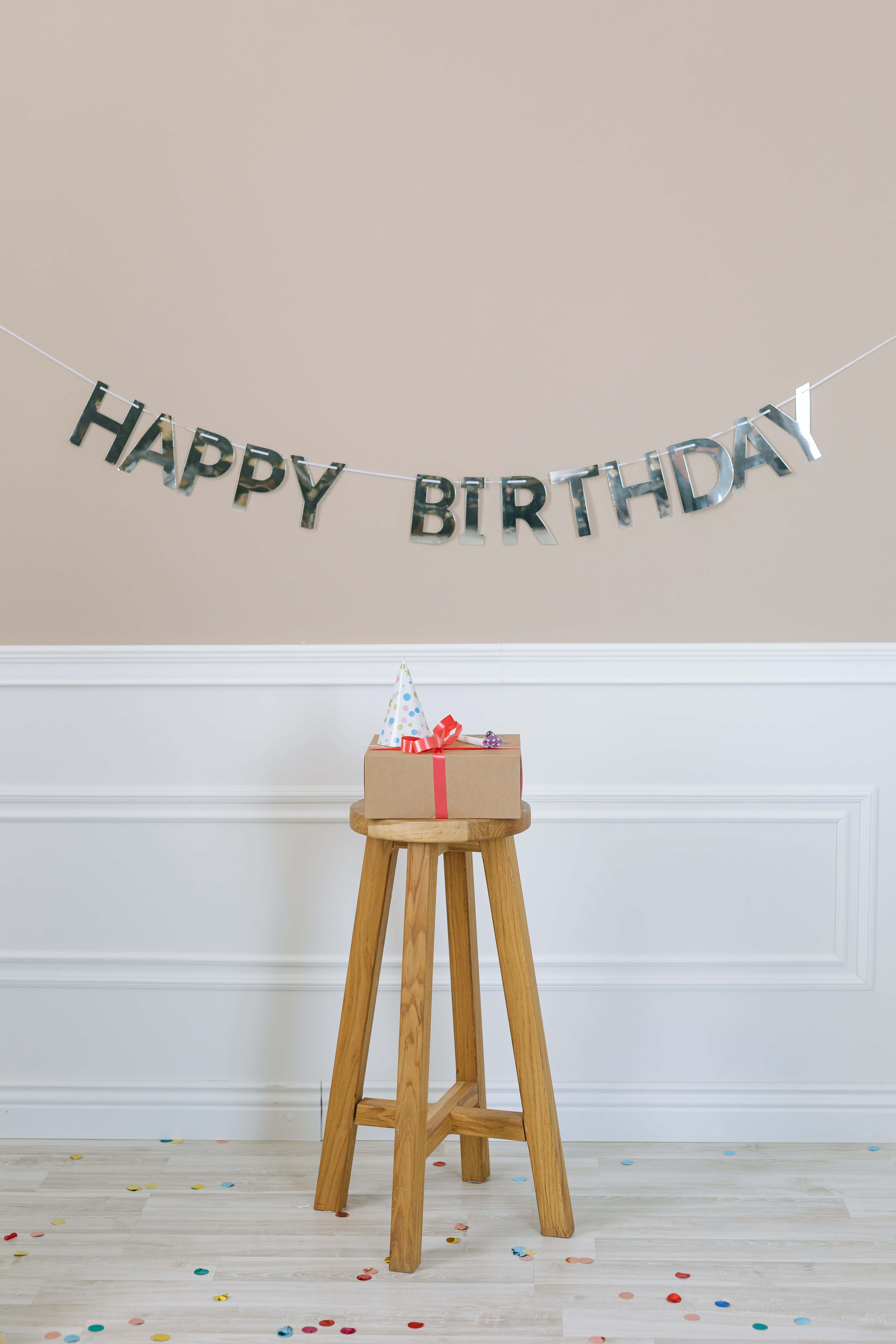 Cute Birthday 桌布 壁紙 — 一個帶有派對帽的生日禮物盒放在木凳上，下方是「生日快樂」橫幅，地上散落著五彩紙屑。