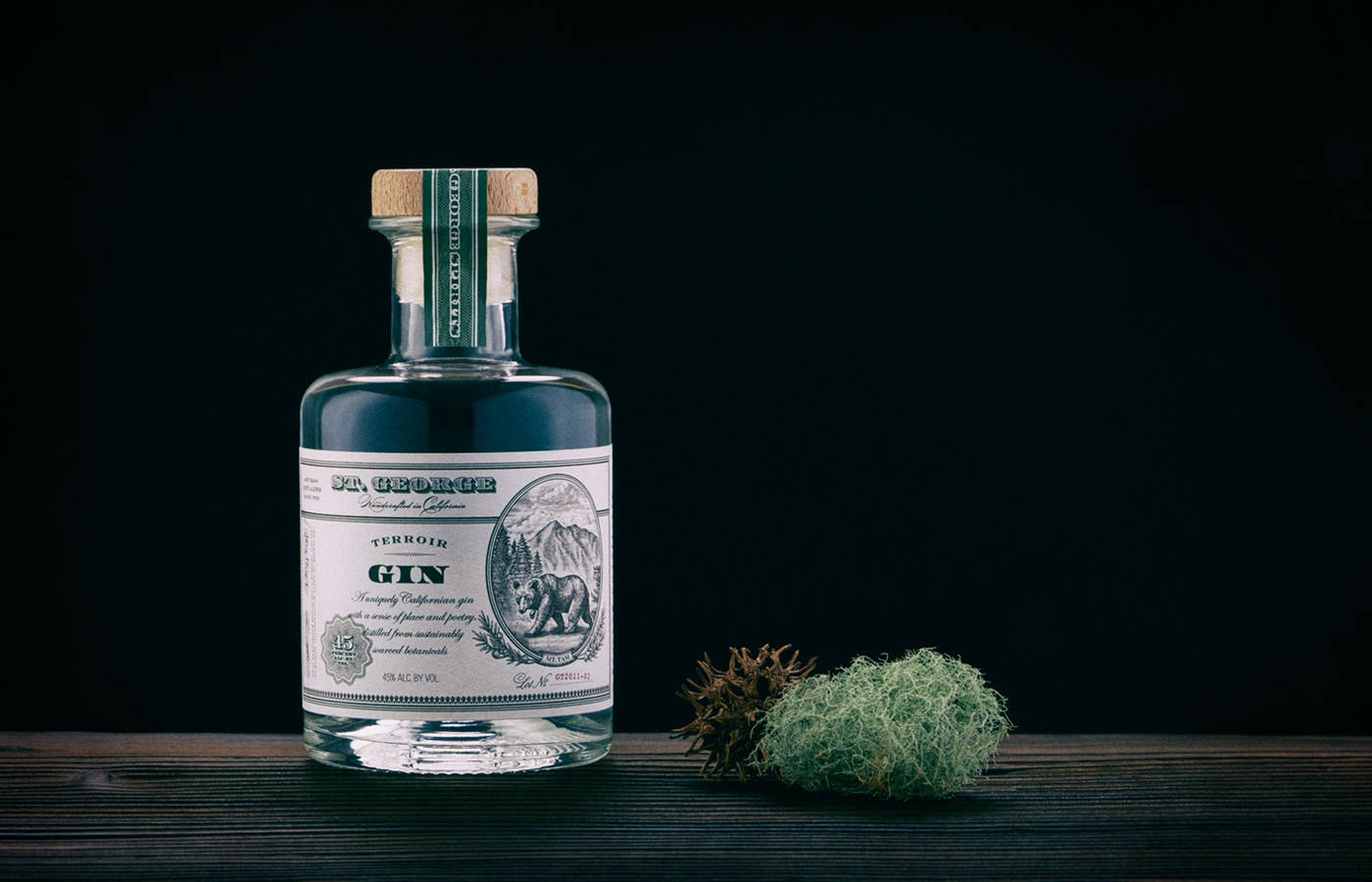 bottle 桌布 壁紙 — 一個帶有熊標籤的透明琴酒瓶放在黑色背景的深色木質表面上，旁邊有苔蘚和松果。