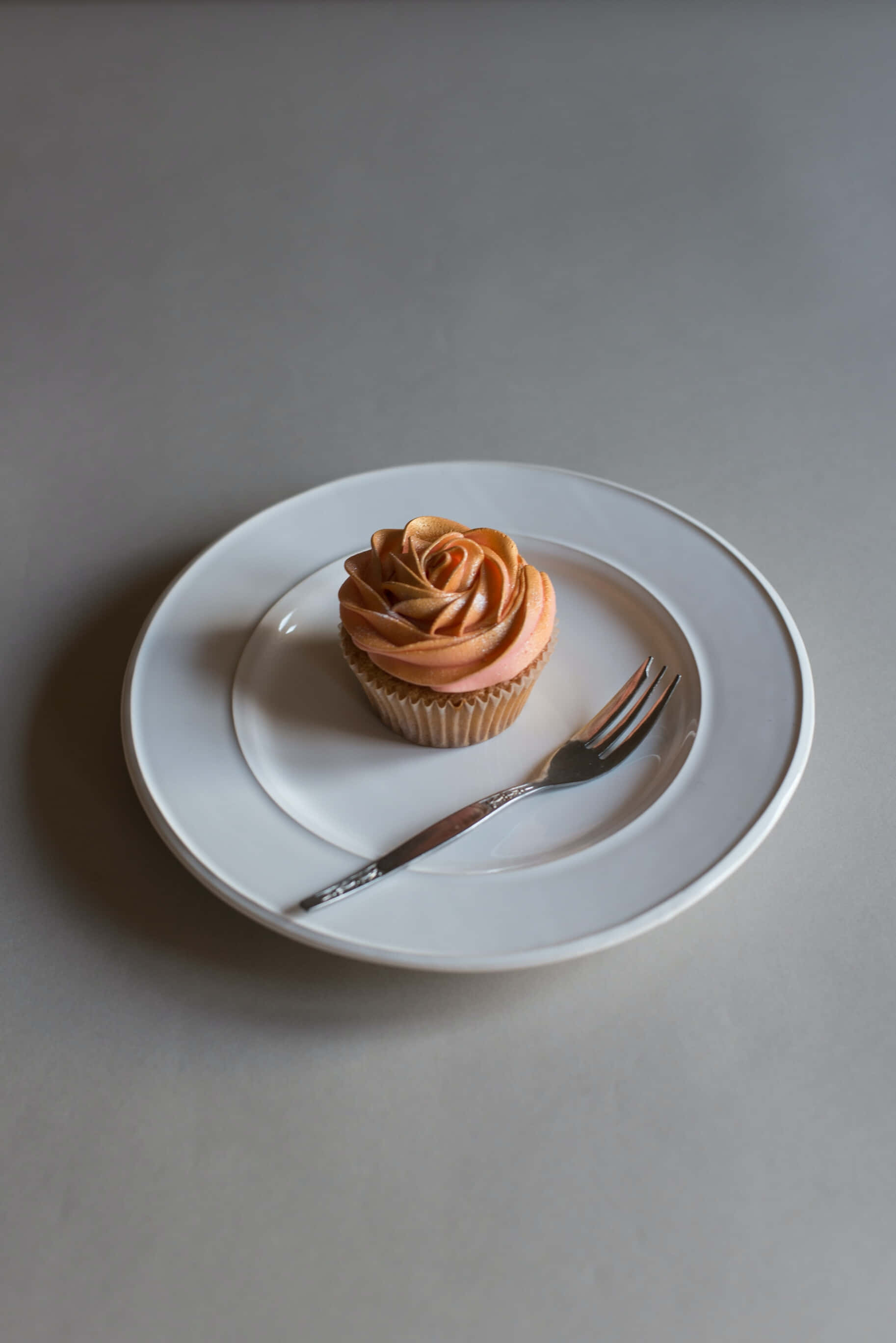 Cupcake 桌布 壁紙 — 一個帶有玫瑰形狀蜜桃糖霜的杯子蛋糕，優雅地放在白色盤子上，旁邊有叉子，背景是簡約灰色。