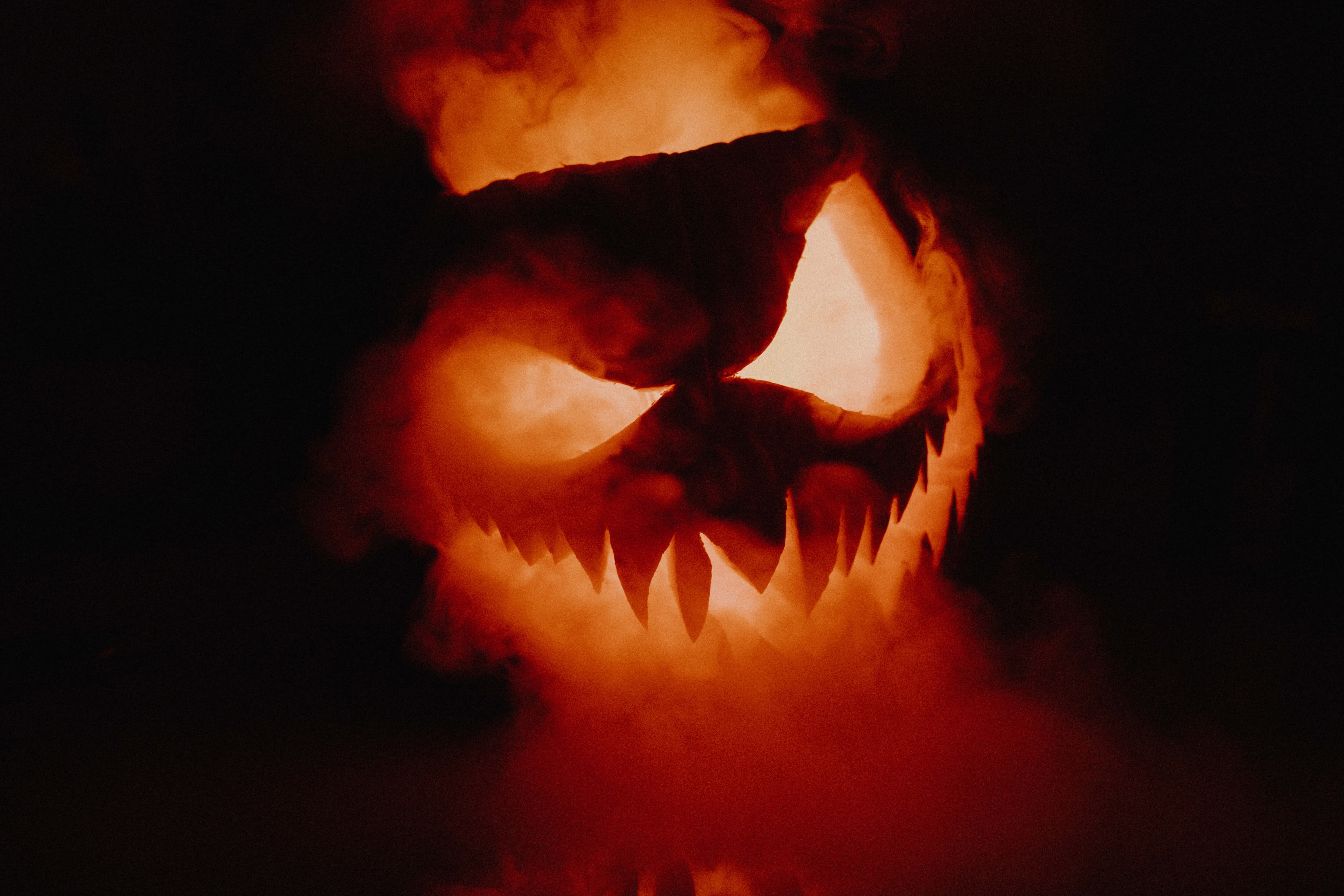Spooky Halloween 桌布 壁紙 — 一個帶有發光橙色眼睛和鋸齒狀牙齒的可怕南瓜燈，在黑色背景的濃煙中悶燒。