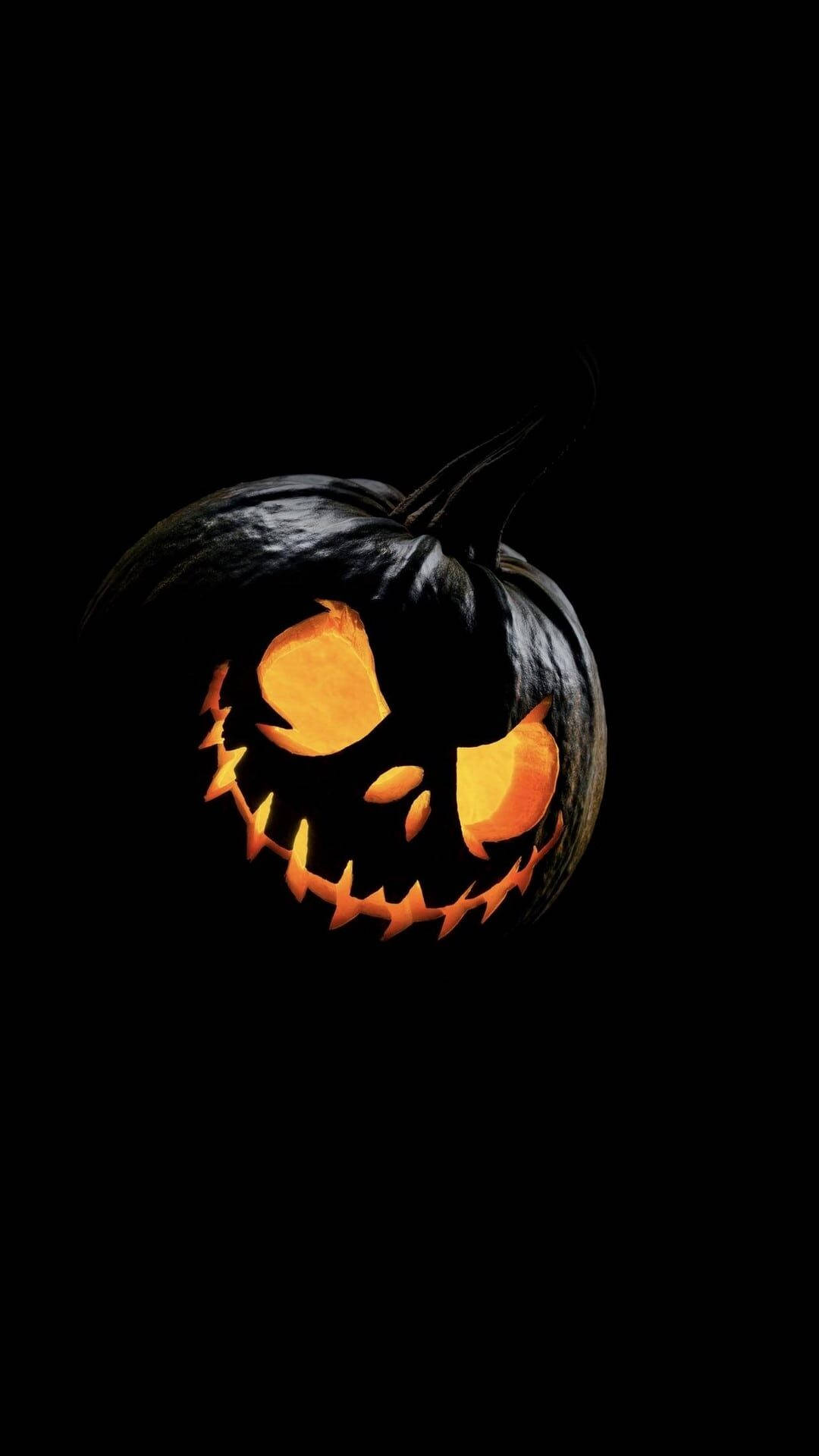 Halloween Phone 桌布 壁紙 — 一個帶有發光橙色眼睛和鋸齒狀牙齒的恐怖南瓜燈，在漆黑的背景下發光。