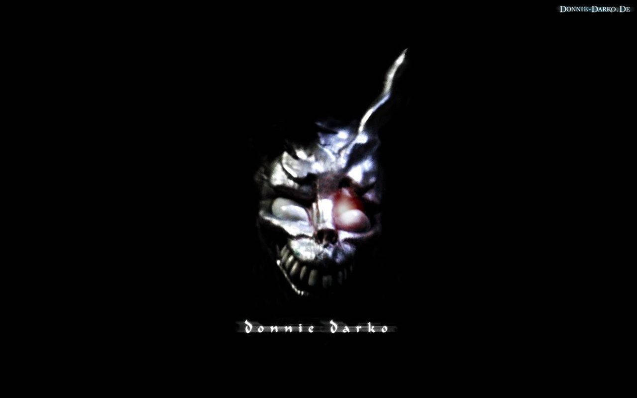 Darko 桌布 壁紙 — 一個帶有發光紅色眼睛和尖銳牙齒的半骷髏半人臉孔，從黑暗中浮現，背景為黑色。