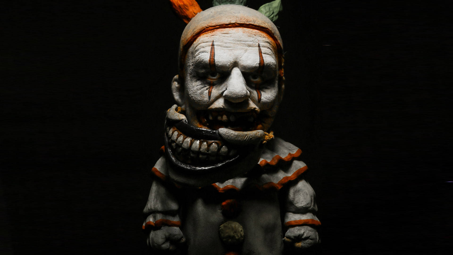 horror villains 桌布 壁紙 — 一個帶有白色塗鴉臉、橙色條紋和咧嘴露齒笑容的可怕小丑雕像，背景為黑色。