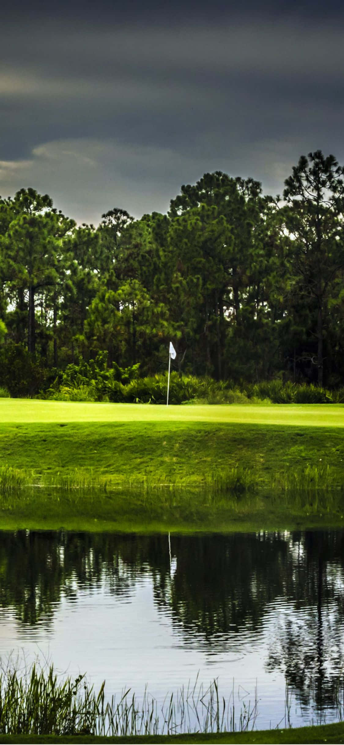 iPhone X Golf Course 桌布 壁紙 — 一個帶有白色旗幟的高爾夫果嶺，矗立在暴風雨天空下，天空倒映在平靜的水障礙中。