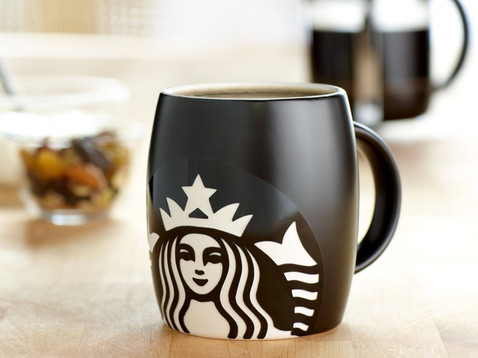 星巴克 桌布 壁紙 — 一個帶有白色標誌的時尚黑色Starbucks馬克杯放在木桌上，旁邊有一碗綜合堅果和一個模糊的咖啡壺。