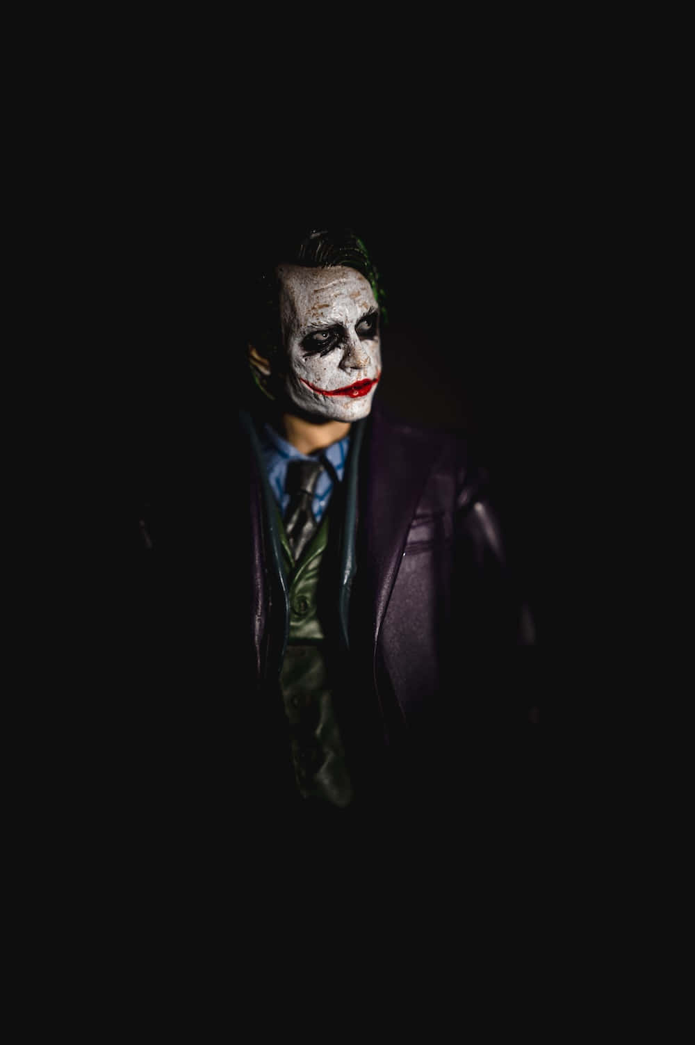Dark Joker 桌布 壁紙 — 一個帶有白色臉部彩繪和紅唇的威脅性人物，穿著紫色西裝，從黑暗中凝視。