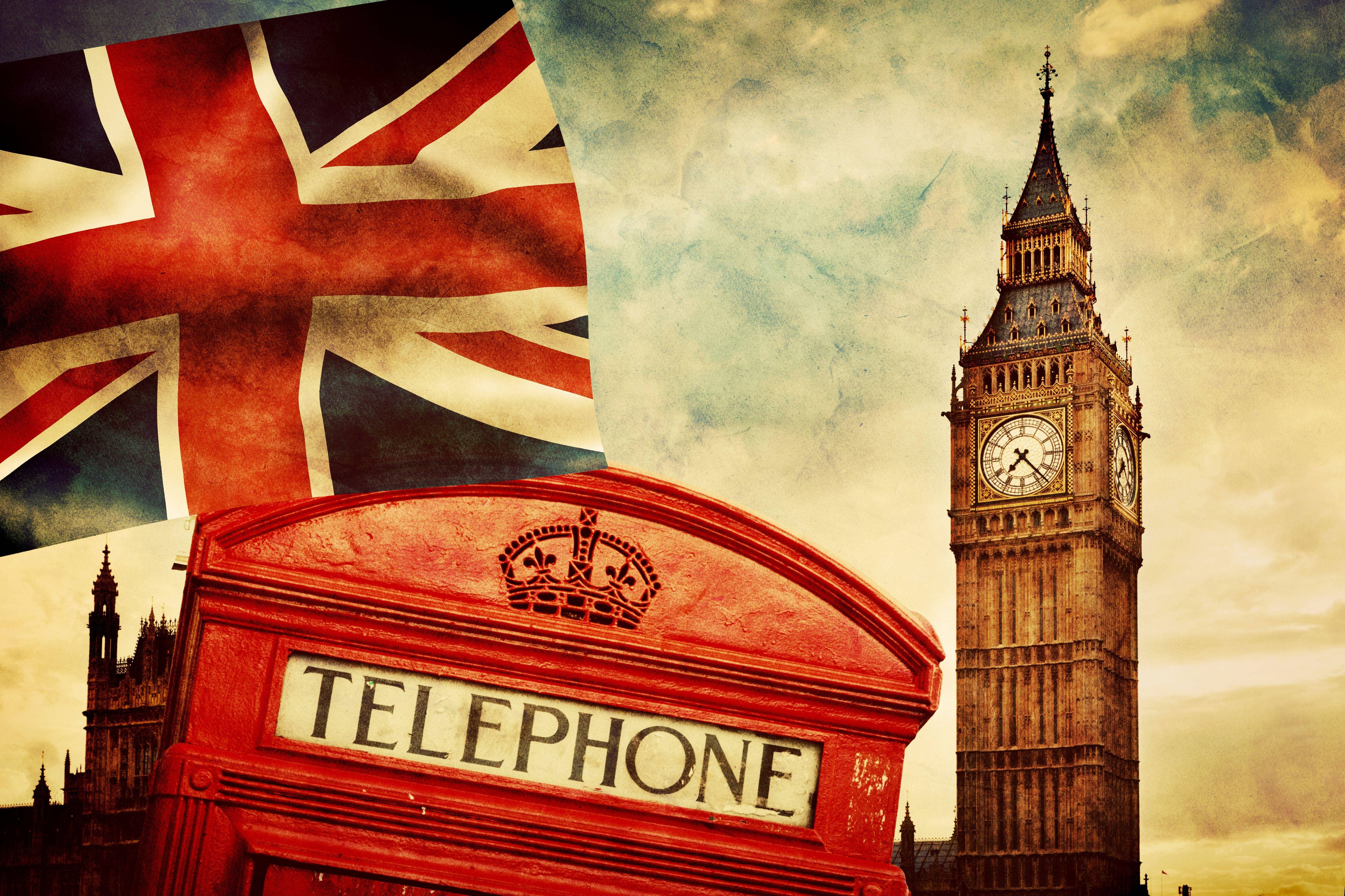 United Kingdom Flag 桌布 壁紙 — 一個帶有皇冠標誌的復古紅色電話亭，矗立在大笨鐘前，背景是戲劇性的多雲天空和一面巨大的英國國旗。