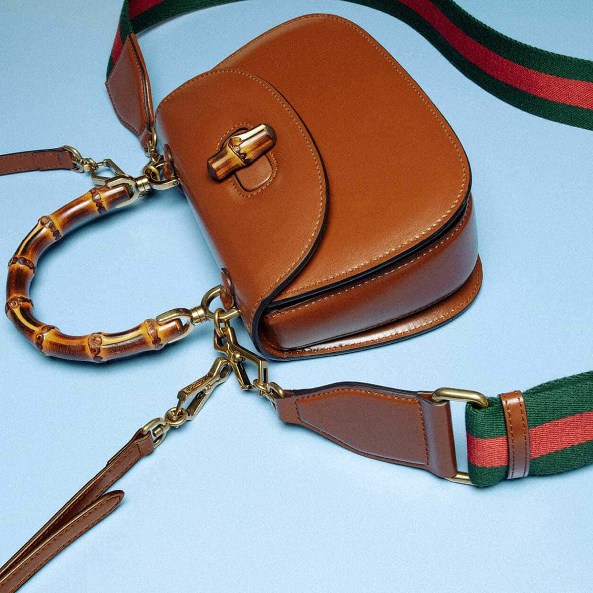 Gucci 桌布 壁紙 — 一個帶有竹手柄和綠色紅色條紋帶的棕褐色皮革手提包放在淺藍色表面上。