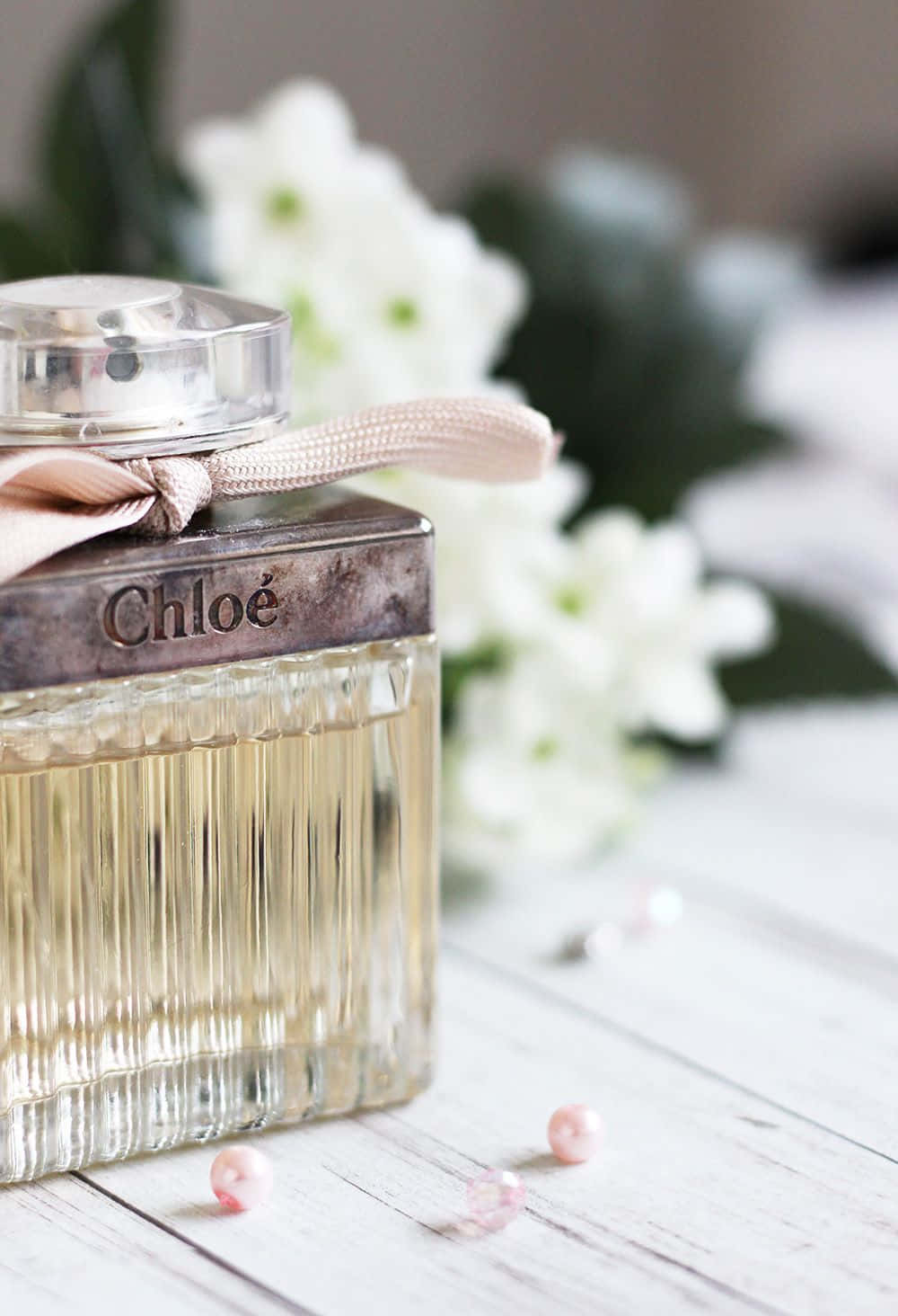 Spring Perfume 桌布 壁紙 — 一個帶有粉紅色絲帶的Chloe香水瓶放在白色木質表面上，周圍環繞著白色花朵和粉紅色珍珠。