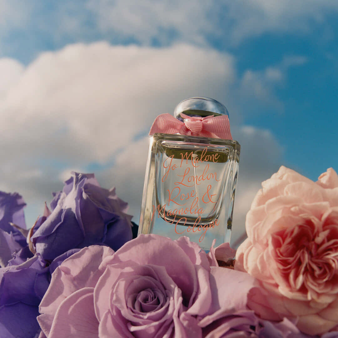 Floral Fragrance 桌布 壁紙 — 一個帶有粉紅色蝴蝶結的Jo Malone香水瓶，放置在茂盛的紫色和粉紅色玫瑰中，背景是明亮的藍天和蓬鬆的雲朵。