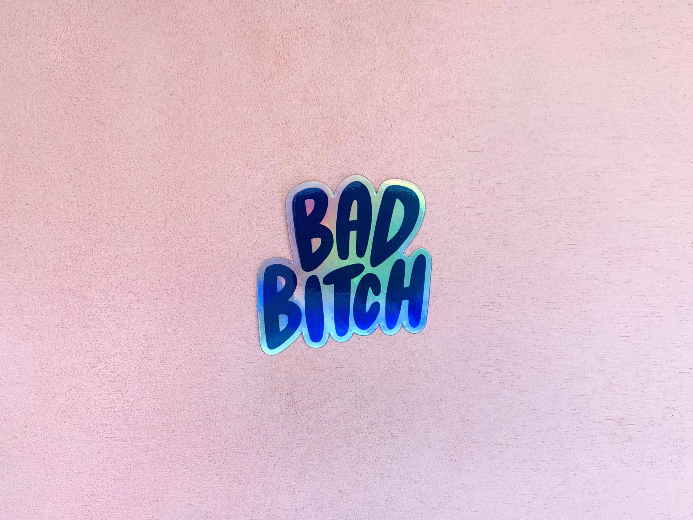 壞女孩 桌布 壁紙 — 一個帶有粗體文字「BAD BITCH」的全息藍色貼紙，居中放置在柔軟的粉紅色紋理表面上。