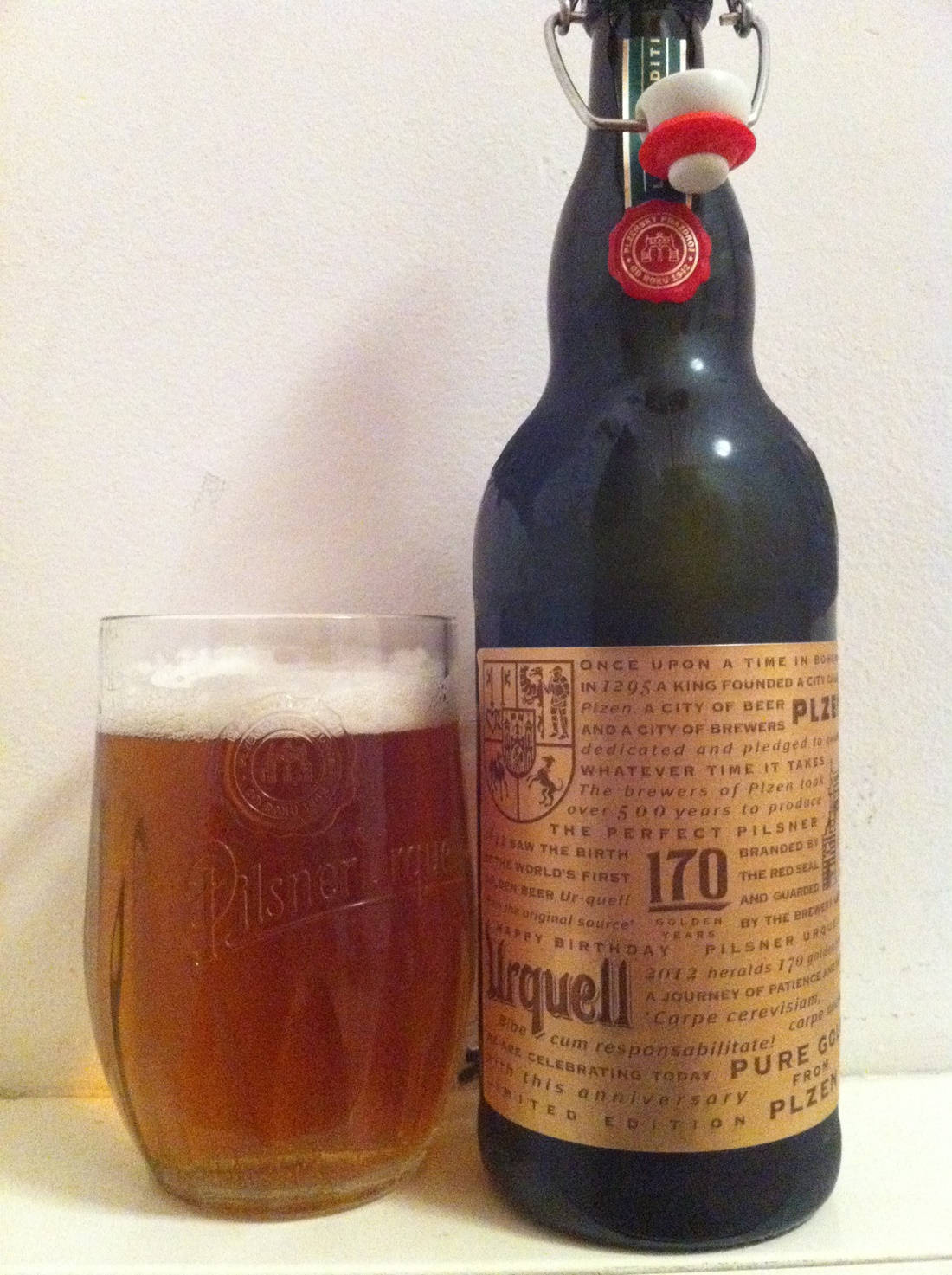 mug 桌布 壁紙 — 一個帶有紅色封條的深色玻璃瓶Pilsner Urquell啤酒，旁邊放著一杯泡沫豐富的琥珀色啤酒，放在純白色表面上。