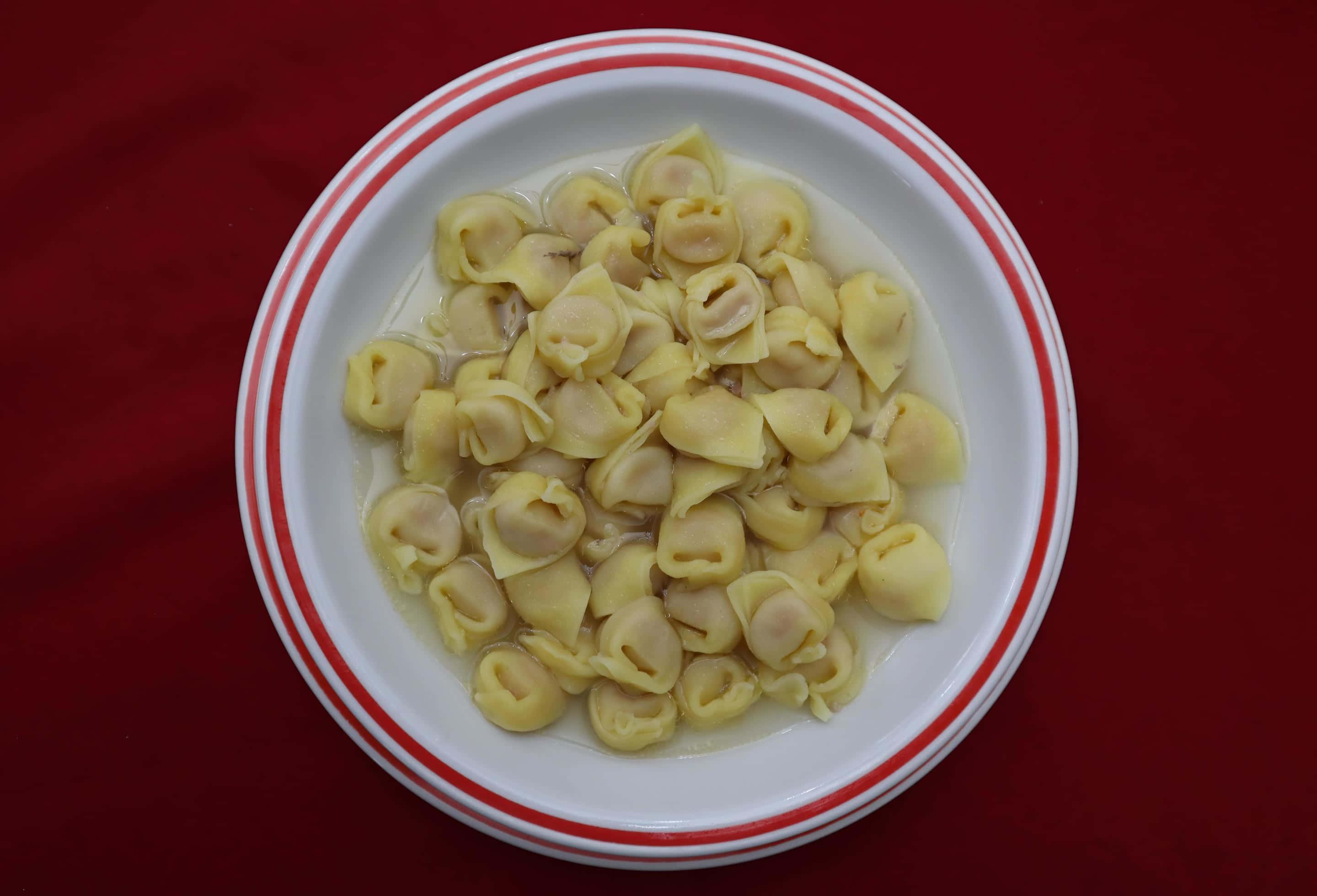 Pasta 桌布 壁紙 — 一個帶有紅色條紋的白色碗，盛載著淺黃色義大利餃在湯中，背景為深紅色。
