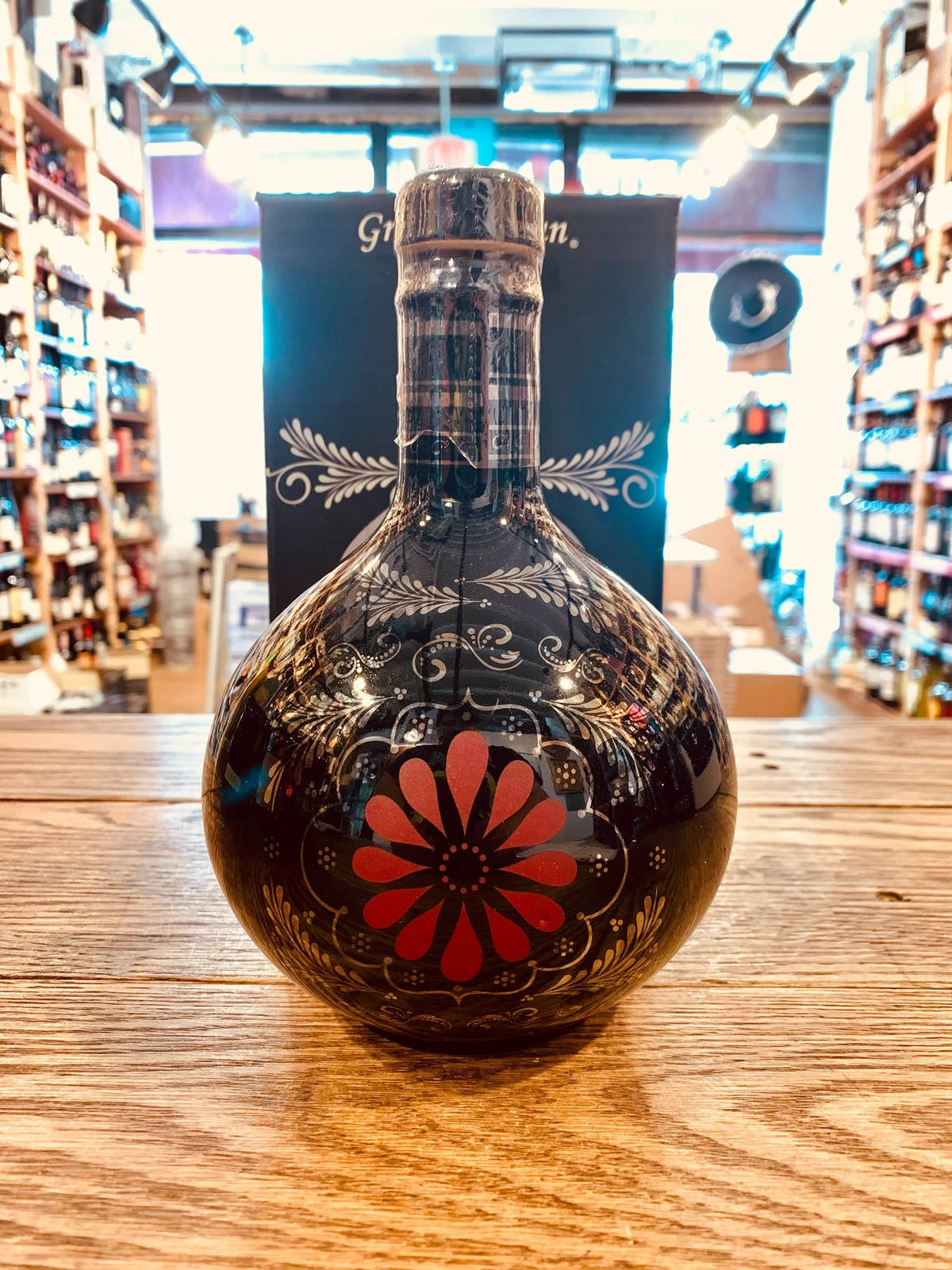 alcohol 桌布 壁紙 — 一個帶有紅色花卉圖案的亮面黑色瓶子放在光線充足的酒類商店的木製櫃檯上。