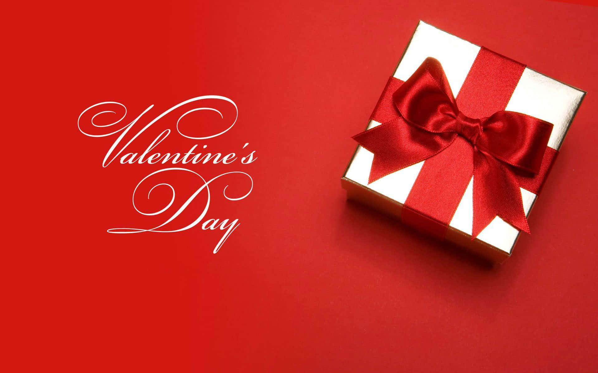 Valentines 桌布 壁紙 — 一個帶有紅色蝴蝶結的白色禮物盒放在純紅色背景上，旁邊是優雅的白色草書文字，寫著Valentine's Day。