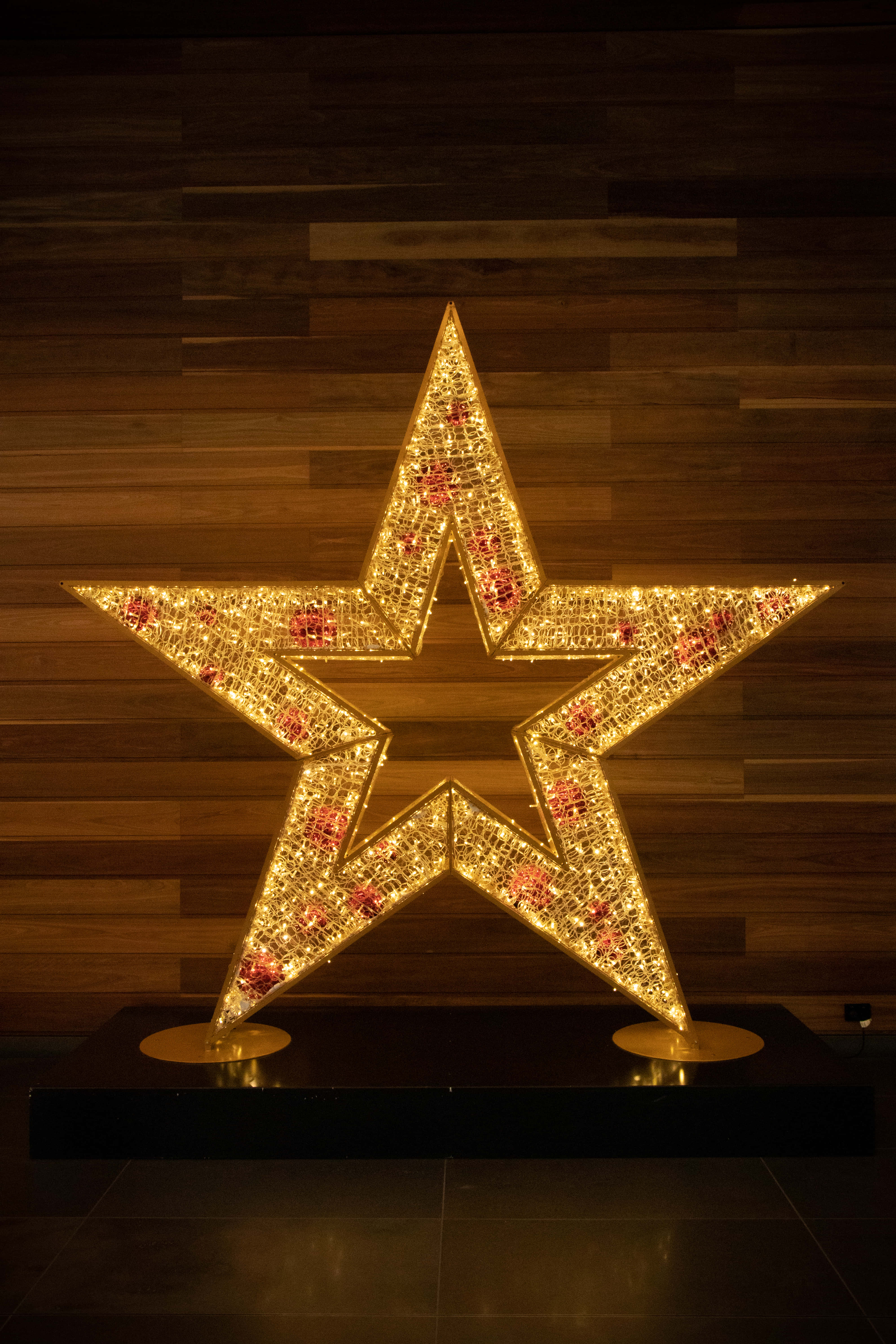 Christmas Star 桌布 壁紙 — 一個帶有紅色點綴的發光金色星星雕塑在深色木牆上被照亮。