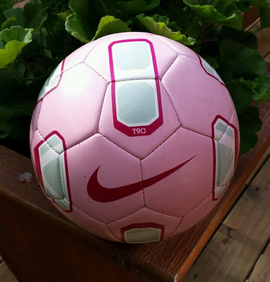 Pink Soccer Ball 桌布 壁紙 — 一個帶有紅色點綴的粉紅色Nike足球放在木質表面上，周圍環繞著茂密的綠葉。