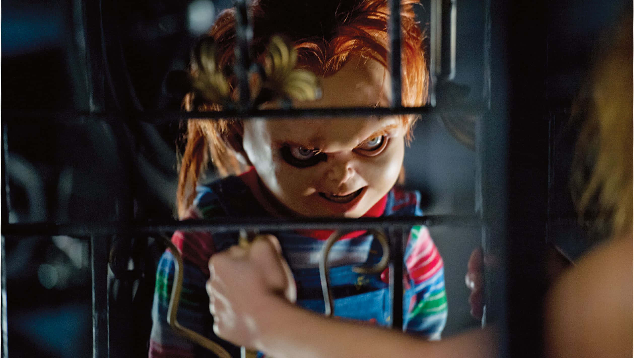 鬼娃恰吉 桌布 壁紙 — 一個帶有紅髮和黑色眼妝的威脅性洋娃娃，透過金屬欄杆窺視著一個人的肩膀。