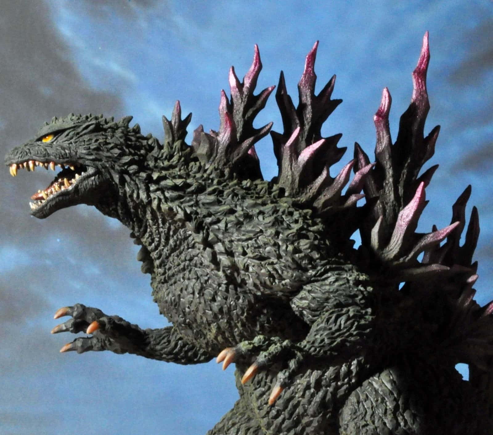 Godzilla 2000 桌布 壁紙 — 一個帶有紋理灰色皮膚和發光黃色眼睛的威脅性哥吉拉公仔，在陰天藍色天空下咆哮。