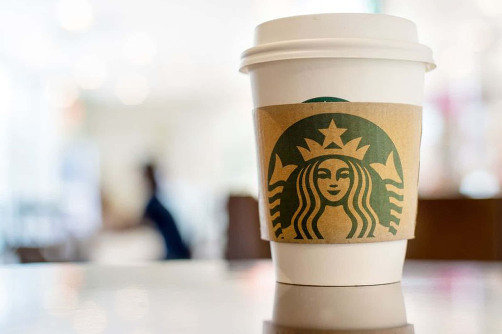 星巴克 桌布 壁紙 — 一個帶有綠色標誌的Starbucks咖啡杯放在明亮咖啡廳的反射表面上，背景模糊。