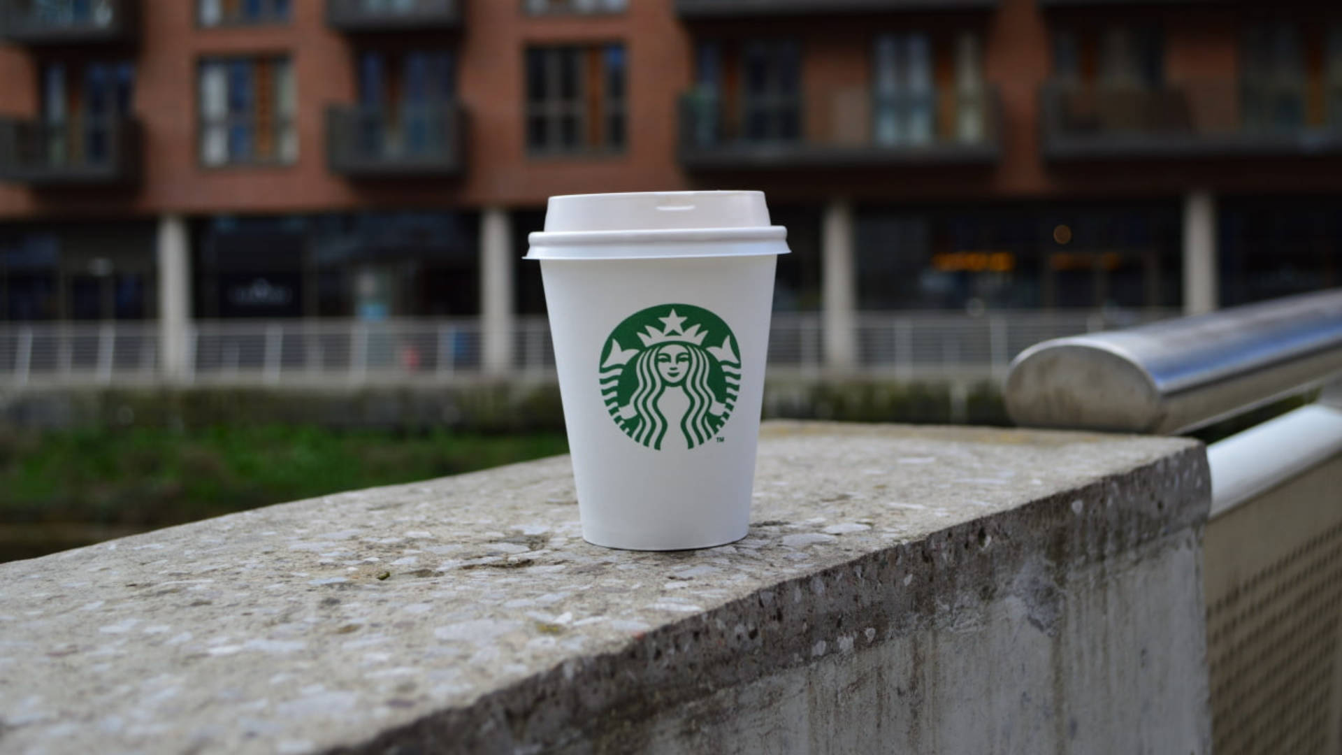 星巴克 桌布 壁紙 — 一個帶有綠色標誌的白色Starbucks咖啡杯放在一個風化的混凝土欄杆上，俯瞰模糊的城市景觀。