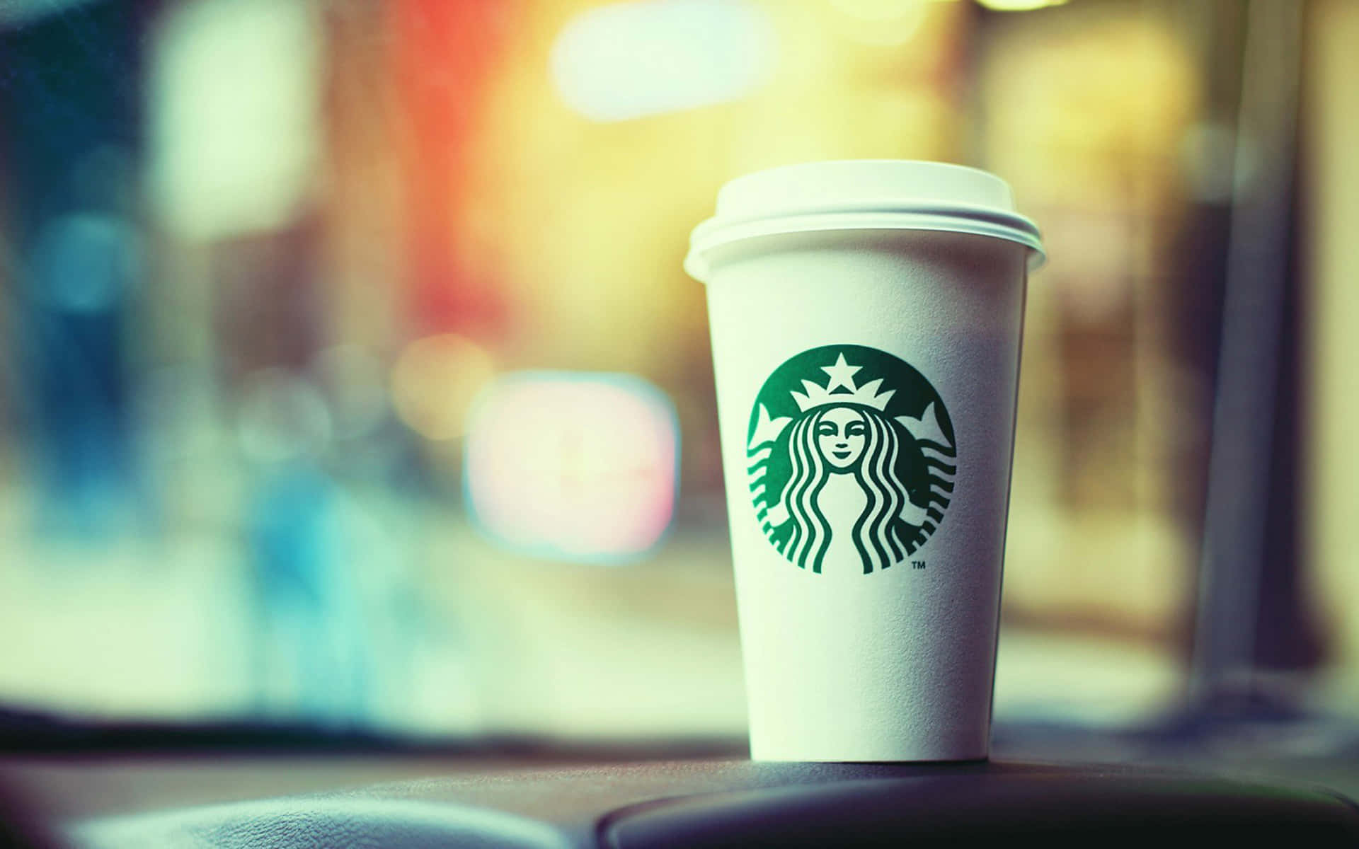 星巴克 桌布 壁紙 — 一個帶有綠色標誌的白色Starbucks咖啡杯放在汽車儀表板上，背景是黃金時刻模糊的城市景觀。
