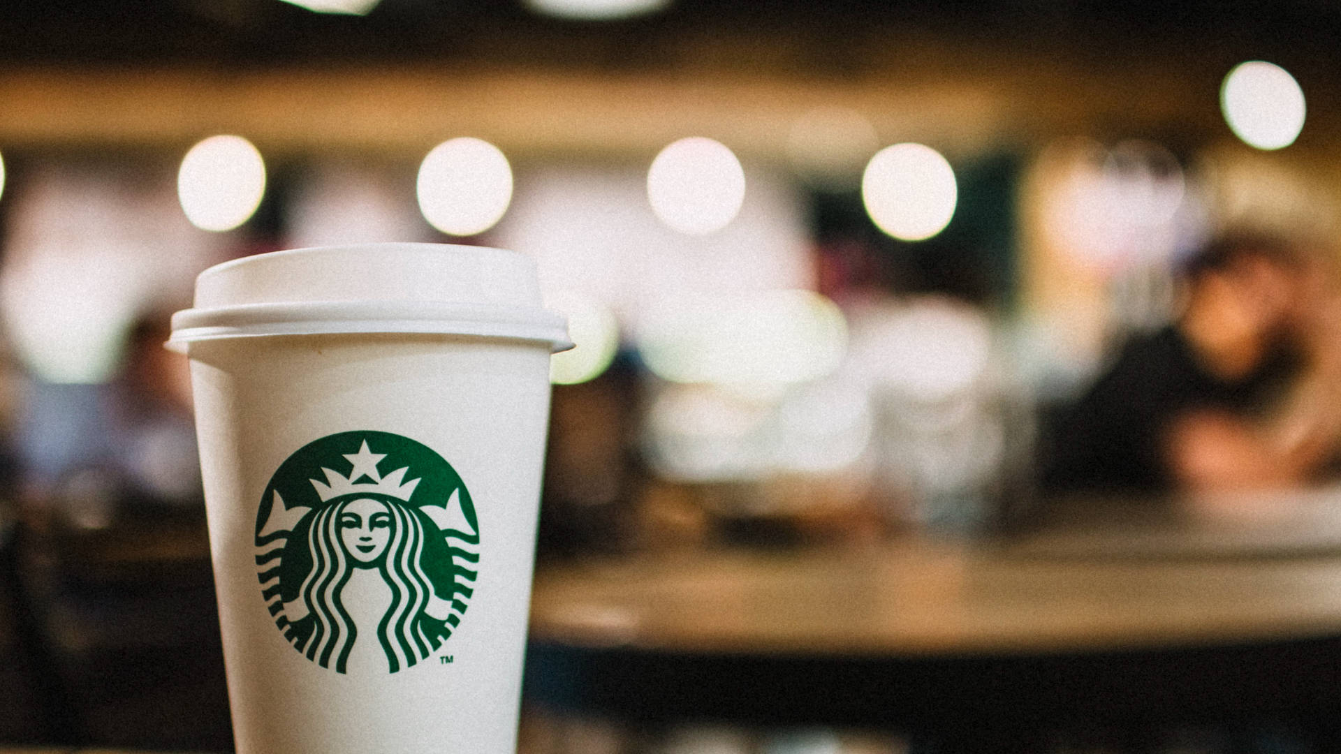 星巴克 桌布 壁紙 — 一個帶有綠色標誌的白色Starbucks咖啡杯，在溫暖模糊的咖啡館背景和散景燈光下清晰對焦。